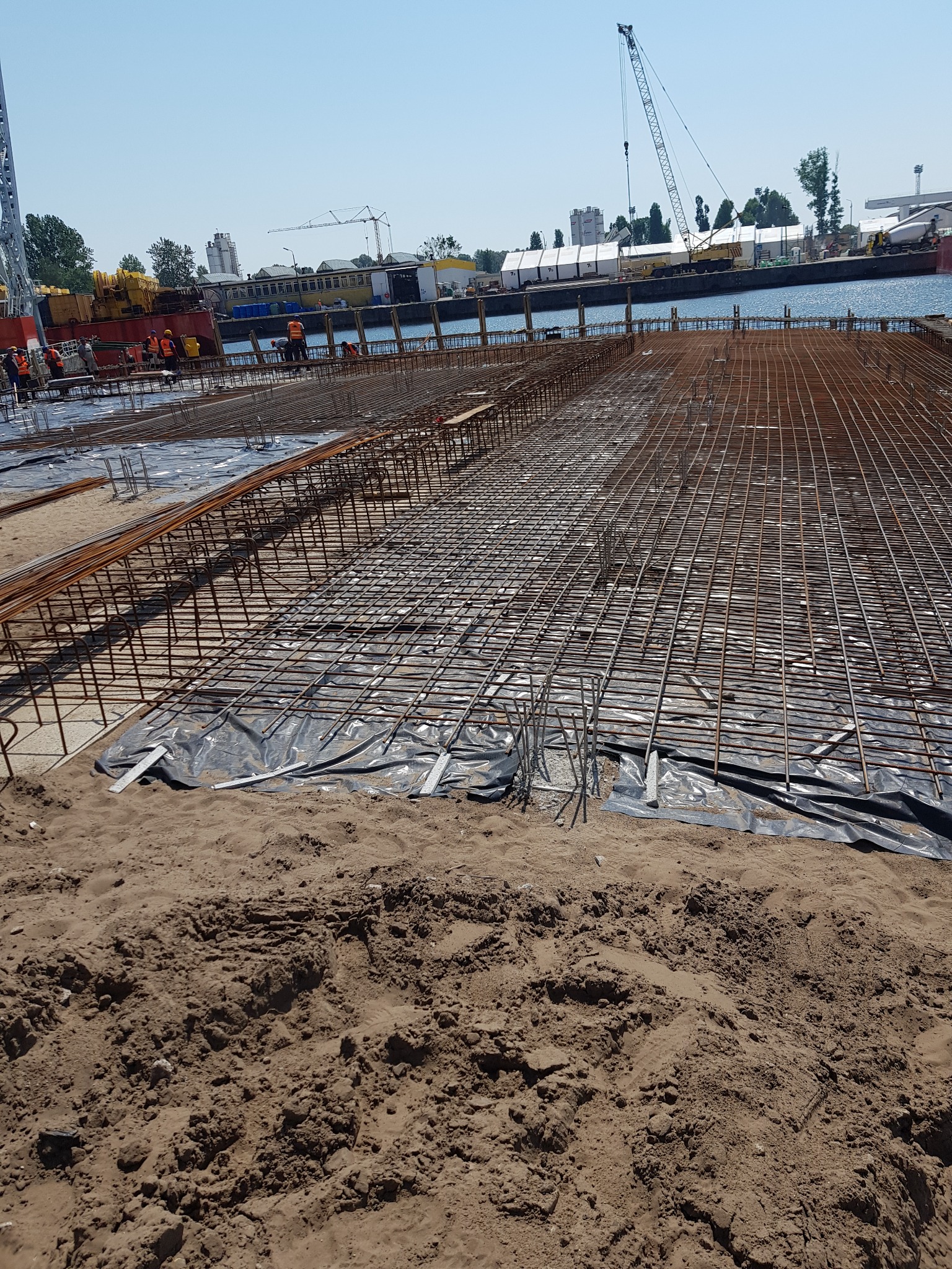 Rozległy szkielet z prętów zbrojeniowych na czarnej folii, przygotowany pod wylewkę fundamentów, z widocznymi pracownikami w pomarańczowych kamizelkach i żurawiami portowymi w tle.