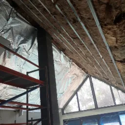 Sufit podwieszany w trakcie montażu: widoczne profile metalowe, izolacja z wełny mineralnej i folia aluminiowa, rusztowanie budowlane oraz okno z widokiem na zieleń.