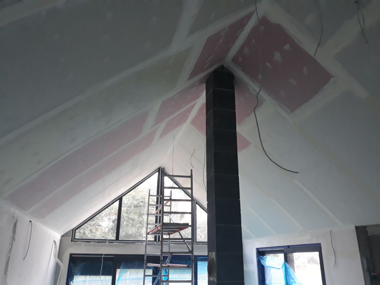 Skośny sufit poddasza w trakcie szpachlowania, widoczne fragmenty płyt gipsowo-kartonowych, czarny komin i aluminiowa drabina, okna zabezpieczone folią.