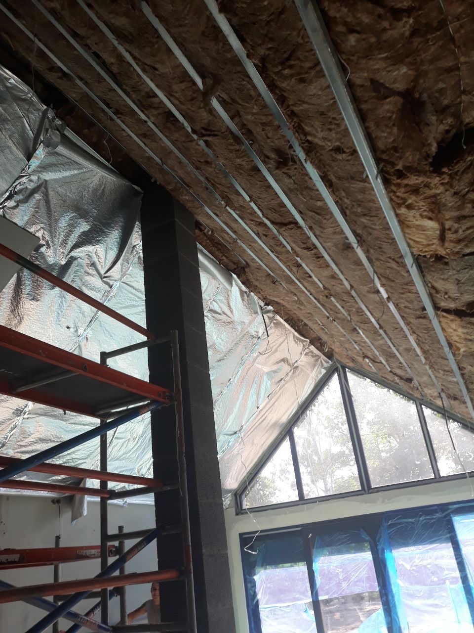 Sufit podwieszany w trakcie montażu: widoczne profile metalowe, izolacja z wełny mineralnej i folia aluminiowa, rusztowanie budowlane oraz okno z widokiem na zieleń.