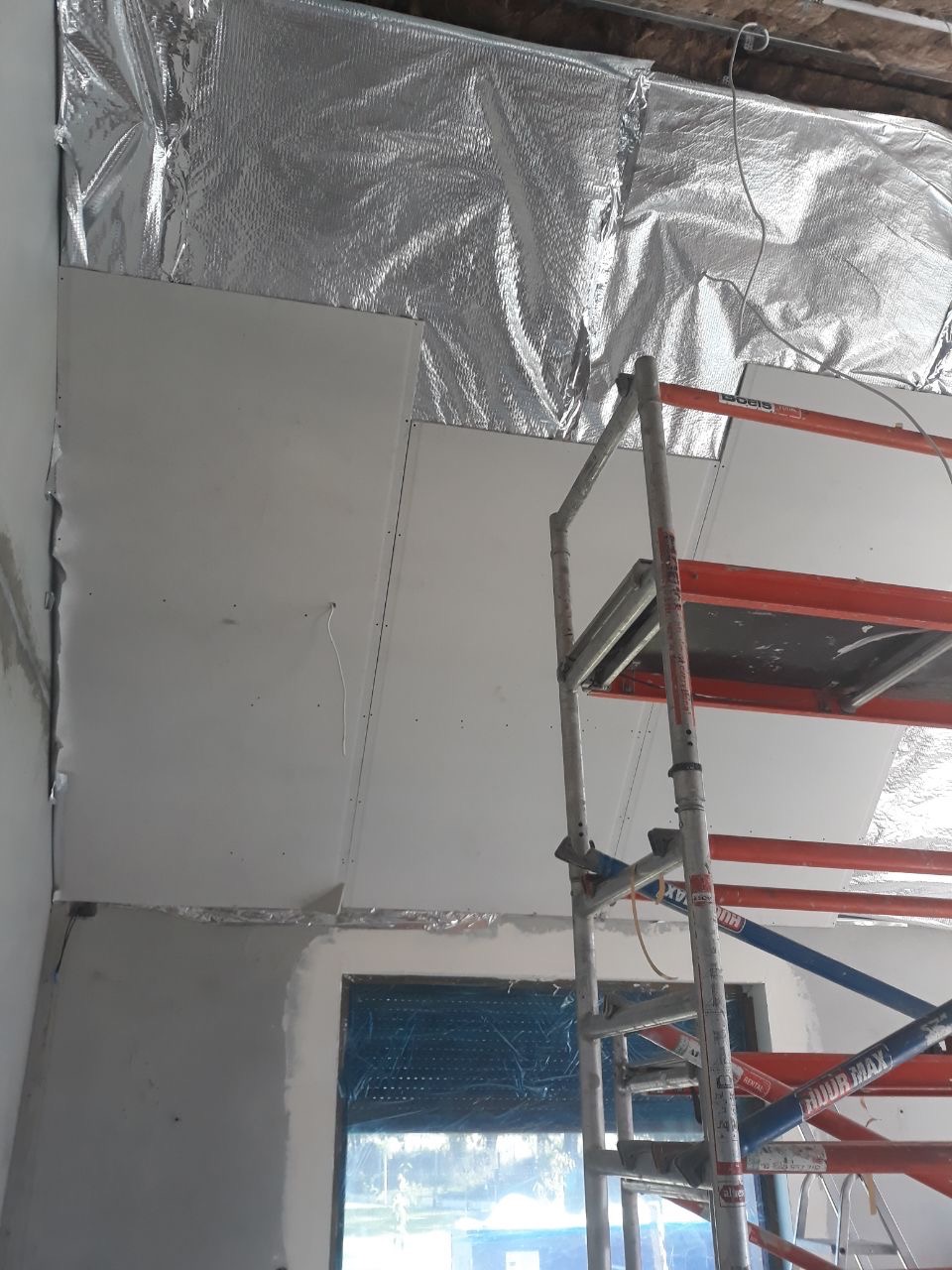 Montaż płyt kartonowo-gipsowych na suficie z izolacją termiczną, widoczne rusztowanie i okno z folią ochronną.