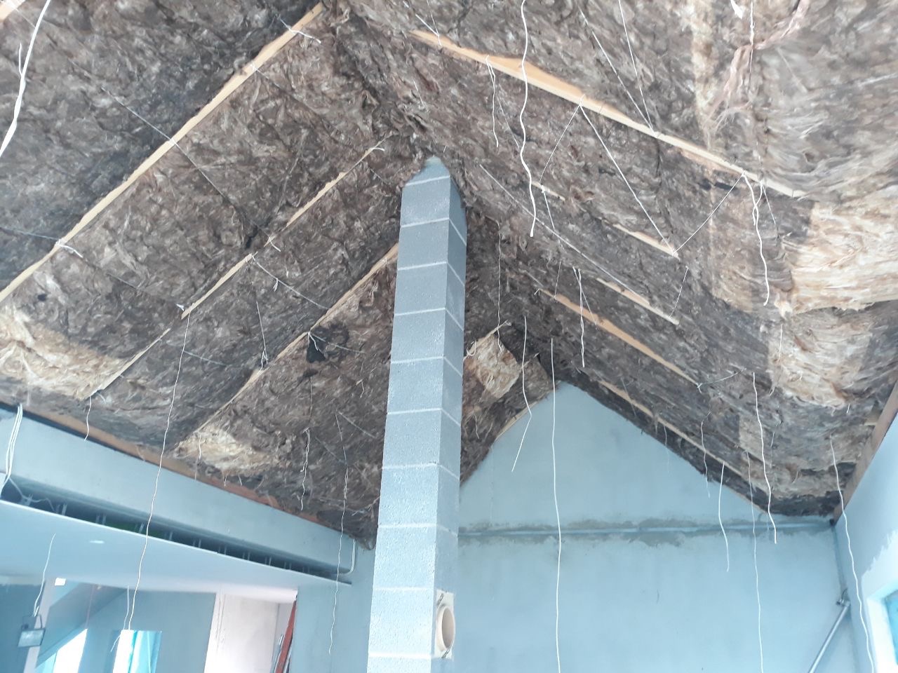 Montaż izolacji termicznej poddasza z widoczną konstrukcją drewnianą i kominem z bloczków betonowych, na ścianach ślady po szpachlowaniu.