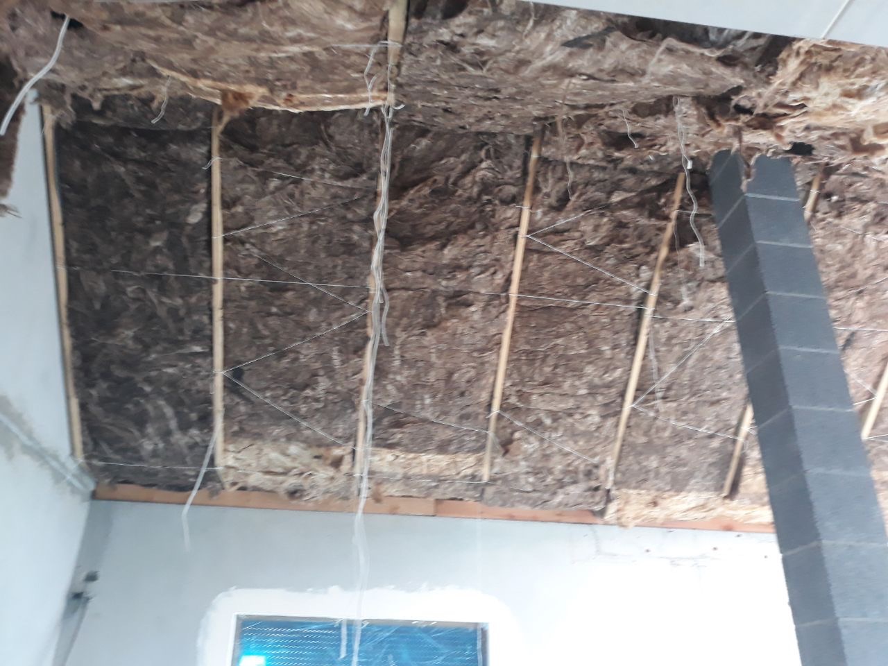 Izolacja poddasza wełną mineralną, widoczne drewniane krokwie i sznurki podtrzymujące materiał, fragment murowanego komina w rogu pomieszczenia.