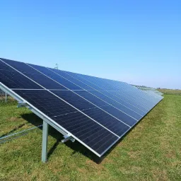 Rząd paneli słonecznych na zielonym trawniku pod błękitnym niebem, widoczne metalowe konstrukcje wsporcze i odległe słupy elektryczne.