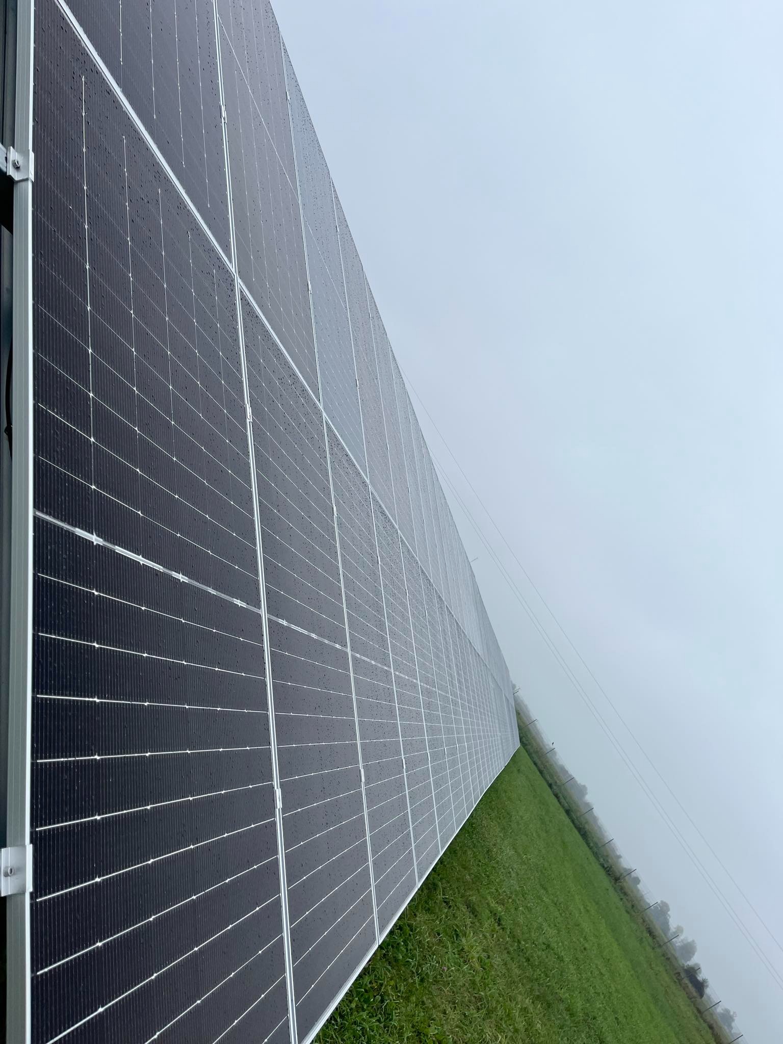 Duży panel fotowoltaiczny z kroplami deszczu na powierzchni, zamontowany na zielonym trawniku w pochmurny dzień.