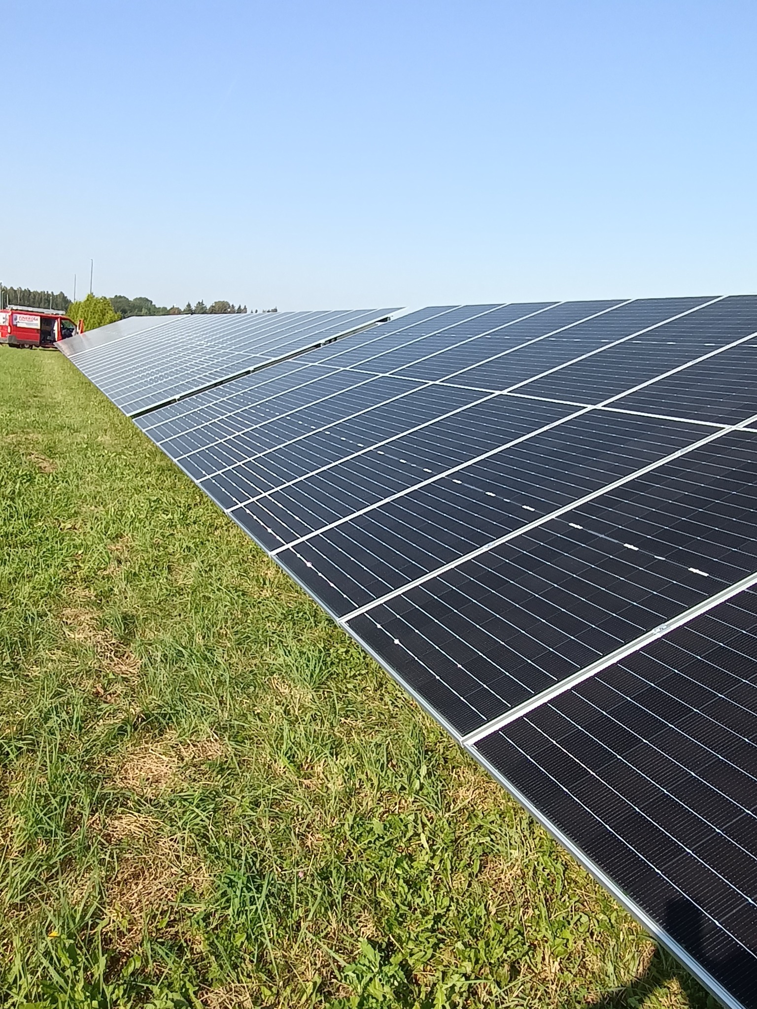 Rozległa instalacja paneli słonecznych na trawiastym terenie, z czerwonym samochodem serwisowym w tle po lewej stronie, widok z niskiej perspektywy.