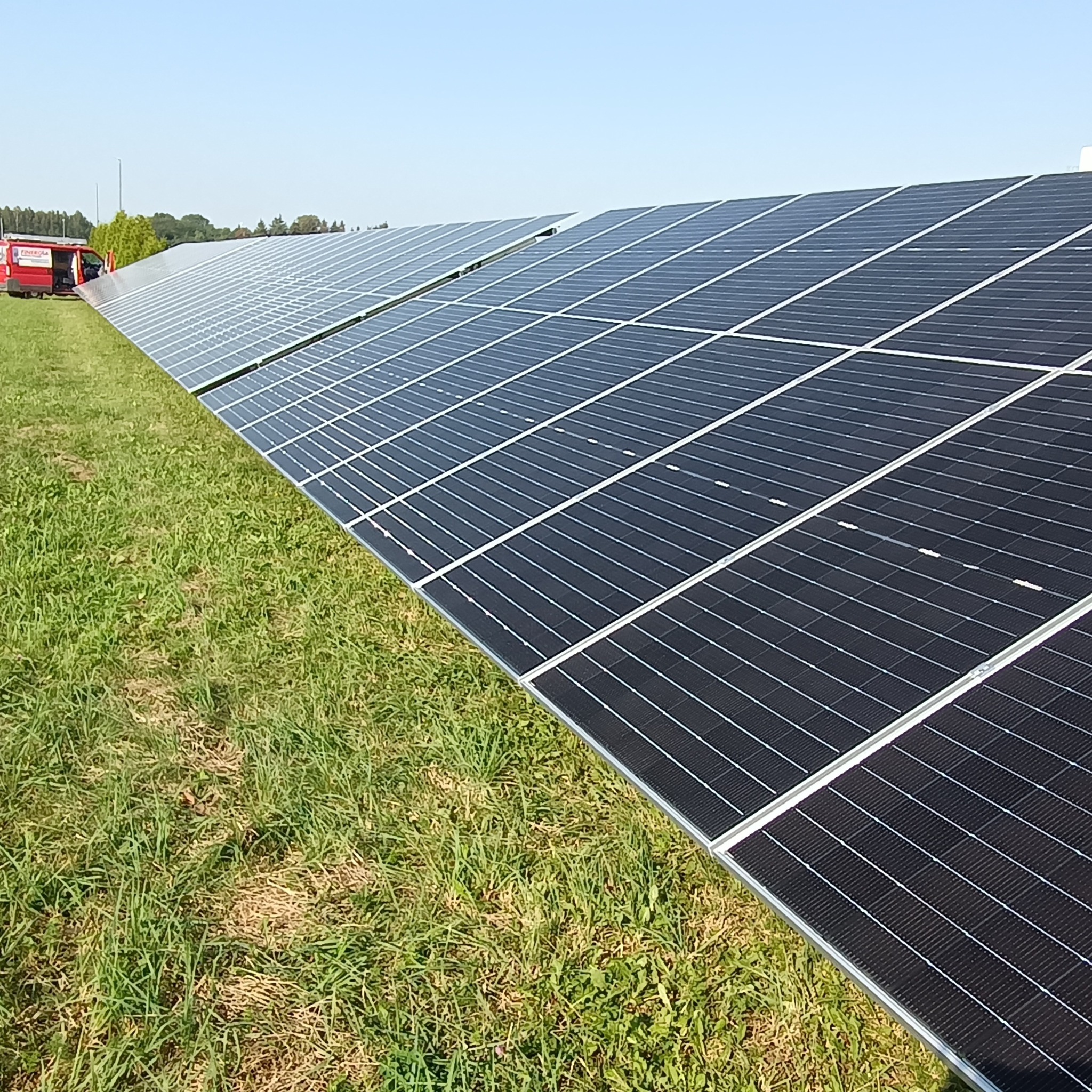 Rząd paneli słonecznych na trawiastym polu, z czerwonym samochodem serwisowym w tle pod błękitnym niebem.