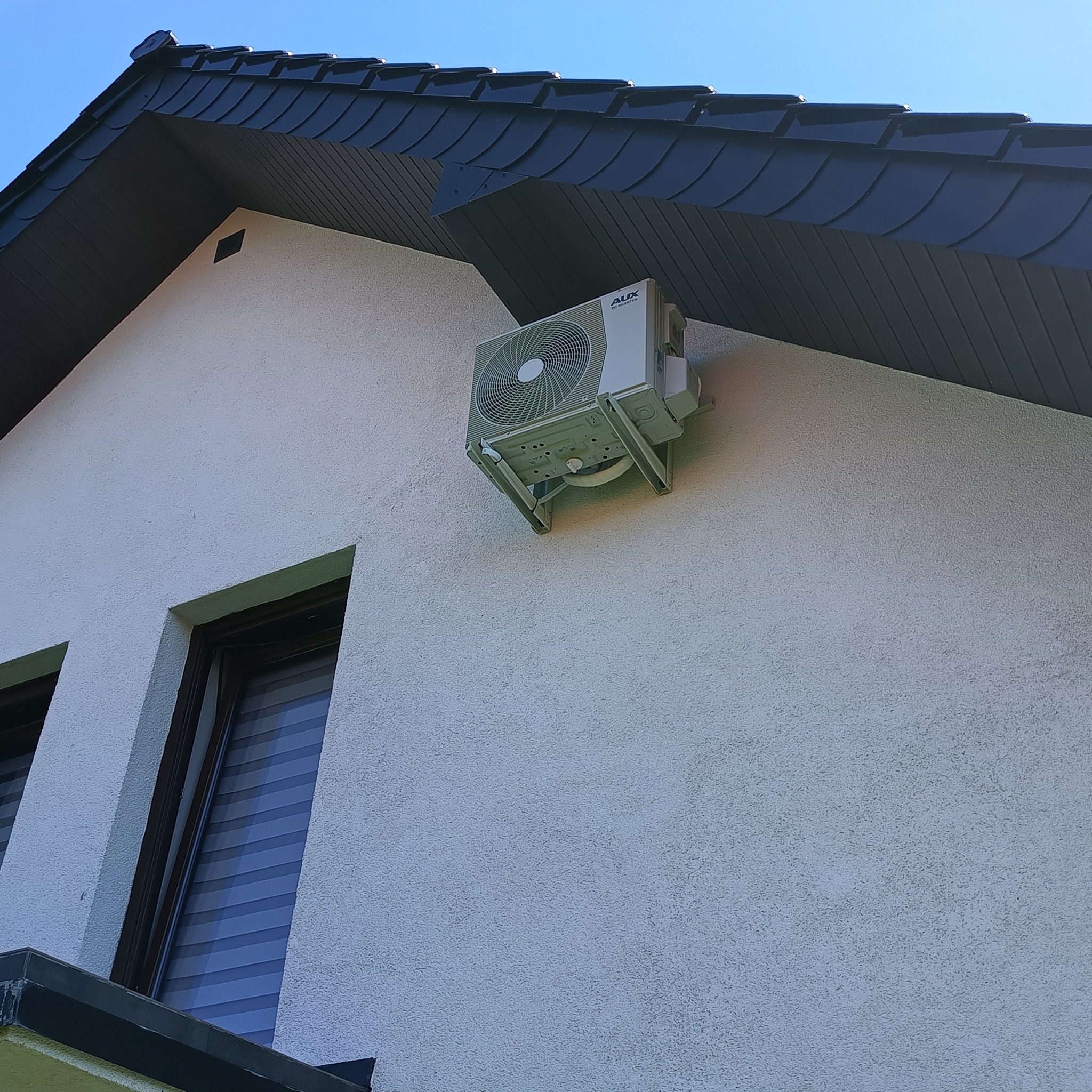 Zewnętrzna jednostka klimatyzacji marki AUX zamontowana na ścianie budynku z fakturą baranka, widoczna z perspektywy żabiej, z fragmentem dachu pokrytego ciemną dachówką w tle.