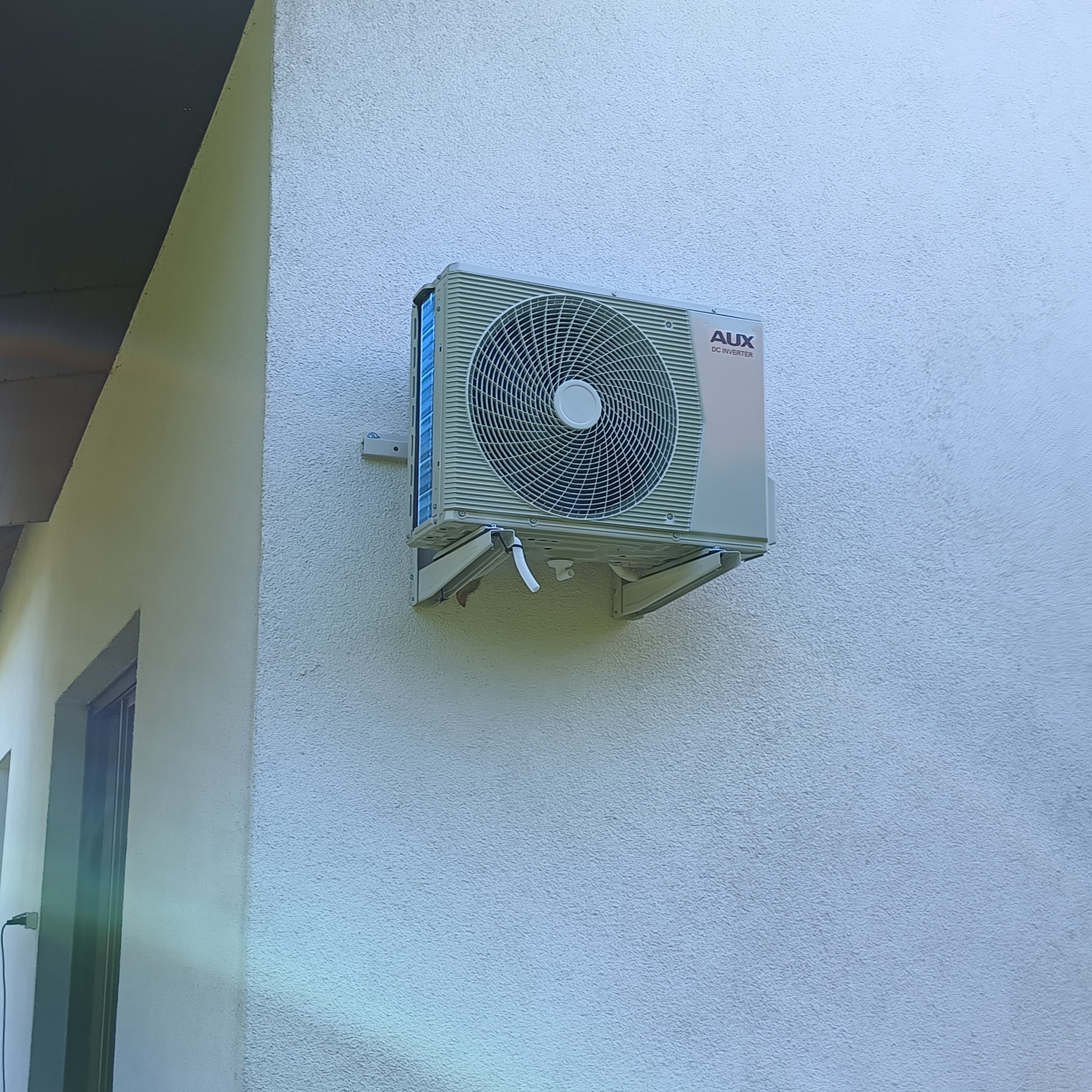 Zewnętrzna jednostka klimatyzacji AUX DC Inverter zamontowana na szarej, teksturowanej ścianie budynku, widoczny fragment okna i dachu.