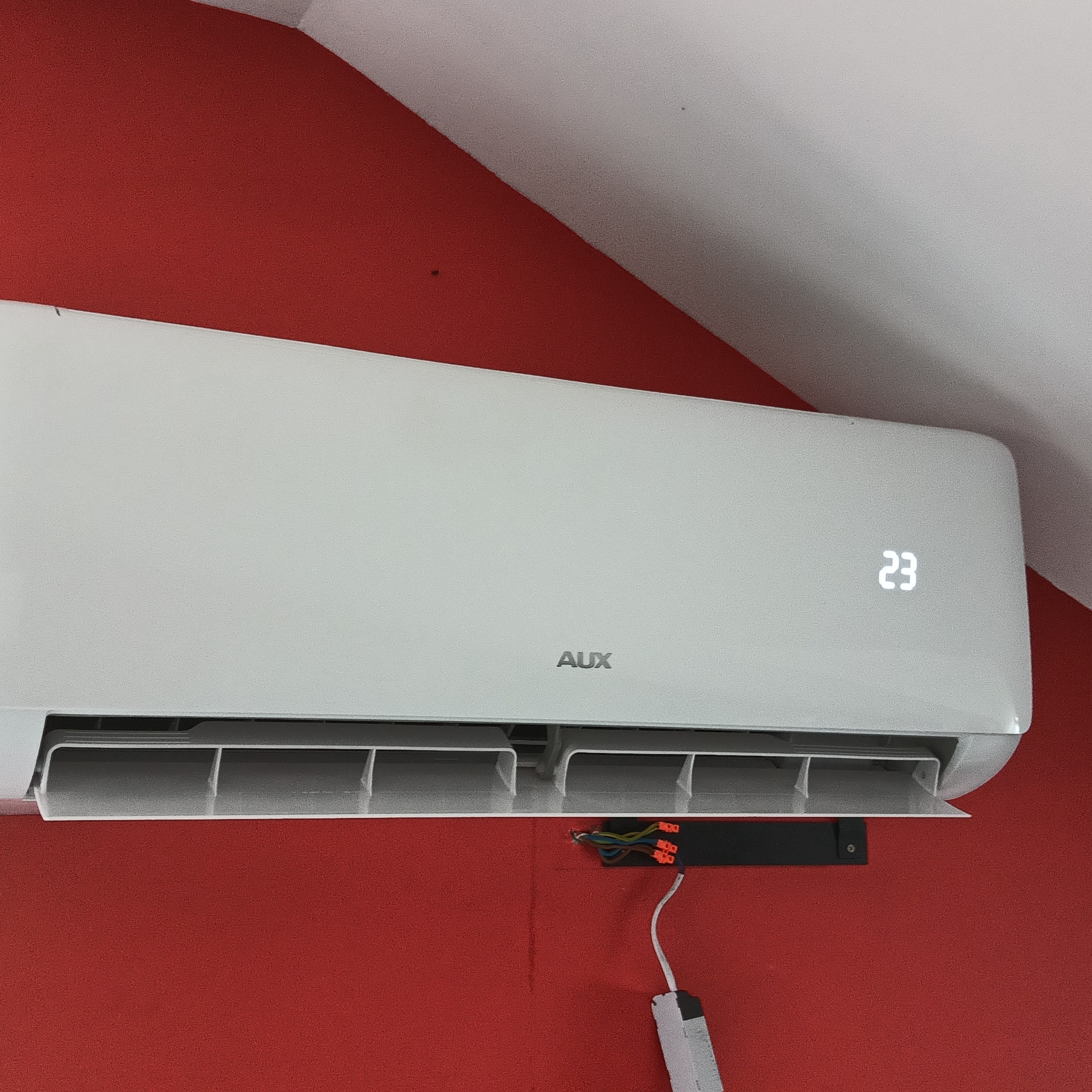 Biały klimatyzator marki AUX zamontowany na czerwonej ścianie, ustawiony na temperaturę 23 stopnie, widoczne podłączenie elektryczne.