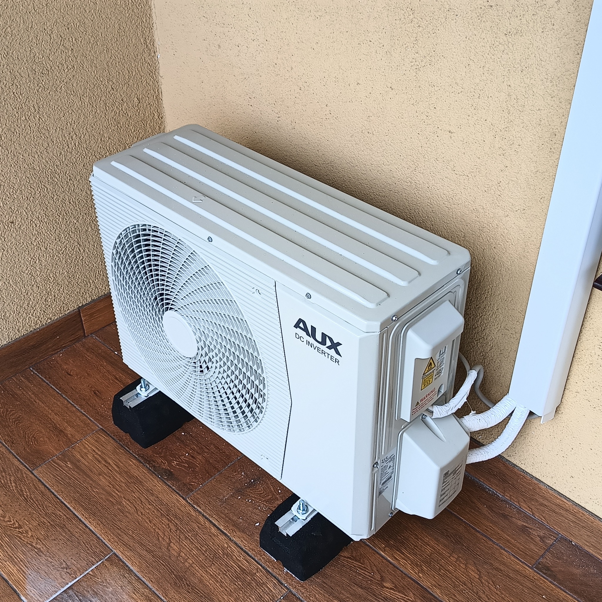 Biała jednostka zewnętrzna klimatyzacji AUX DC Inverter zamontowana na czarnych gumowych podstawkach na drewnianej podłodze, z widocznymi przewodami w białej osłonie, na tle beżowej ściany.