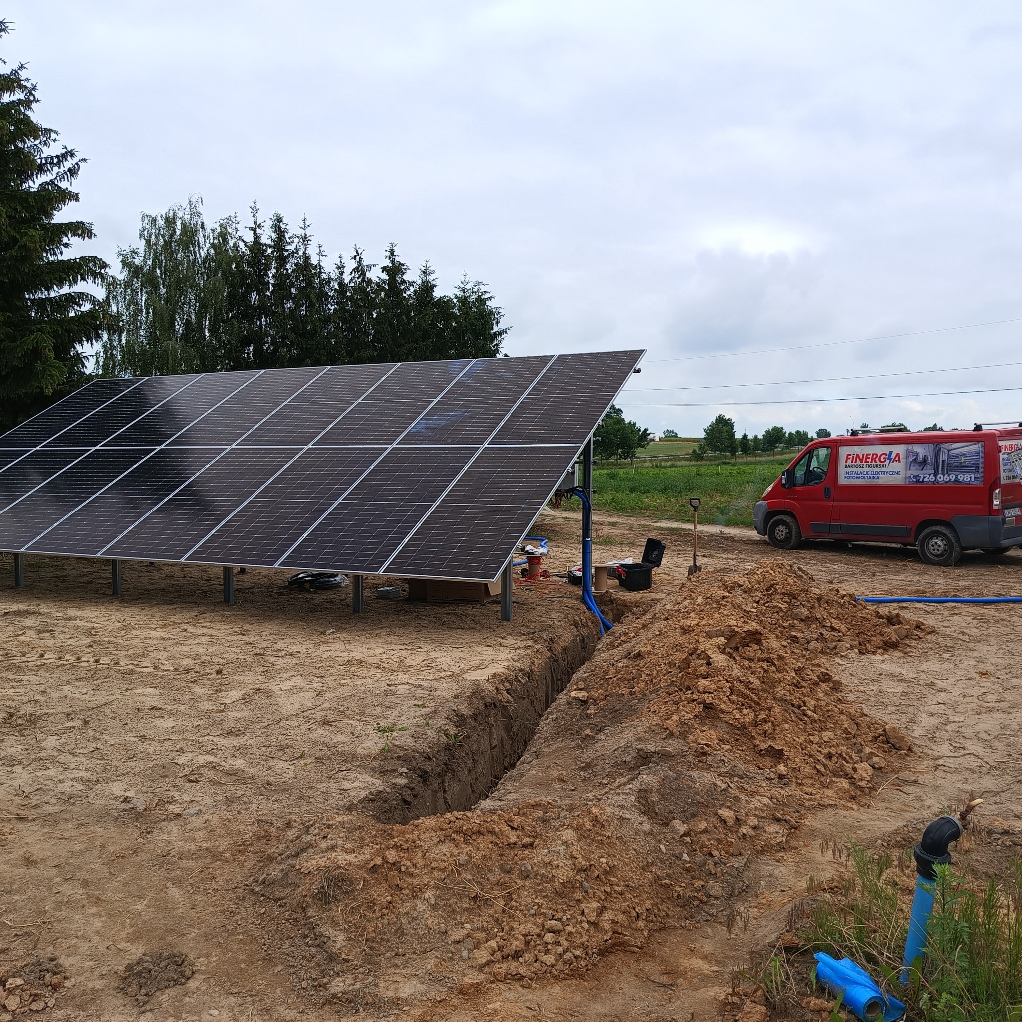 Panel słoneczny na metalowych podporach, częściowo zainstalowany na piaszczystym terenie, z wykopanym rowem na kable i czerwonym vanem firmy z reklamą instalacji elektrycznych i fotowoltaicznych...