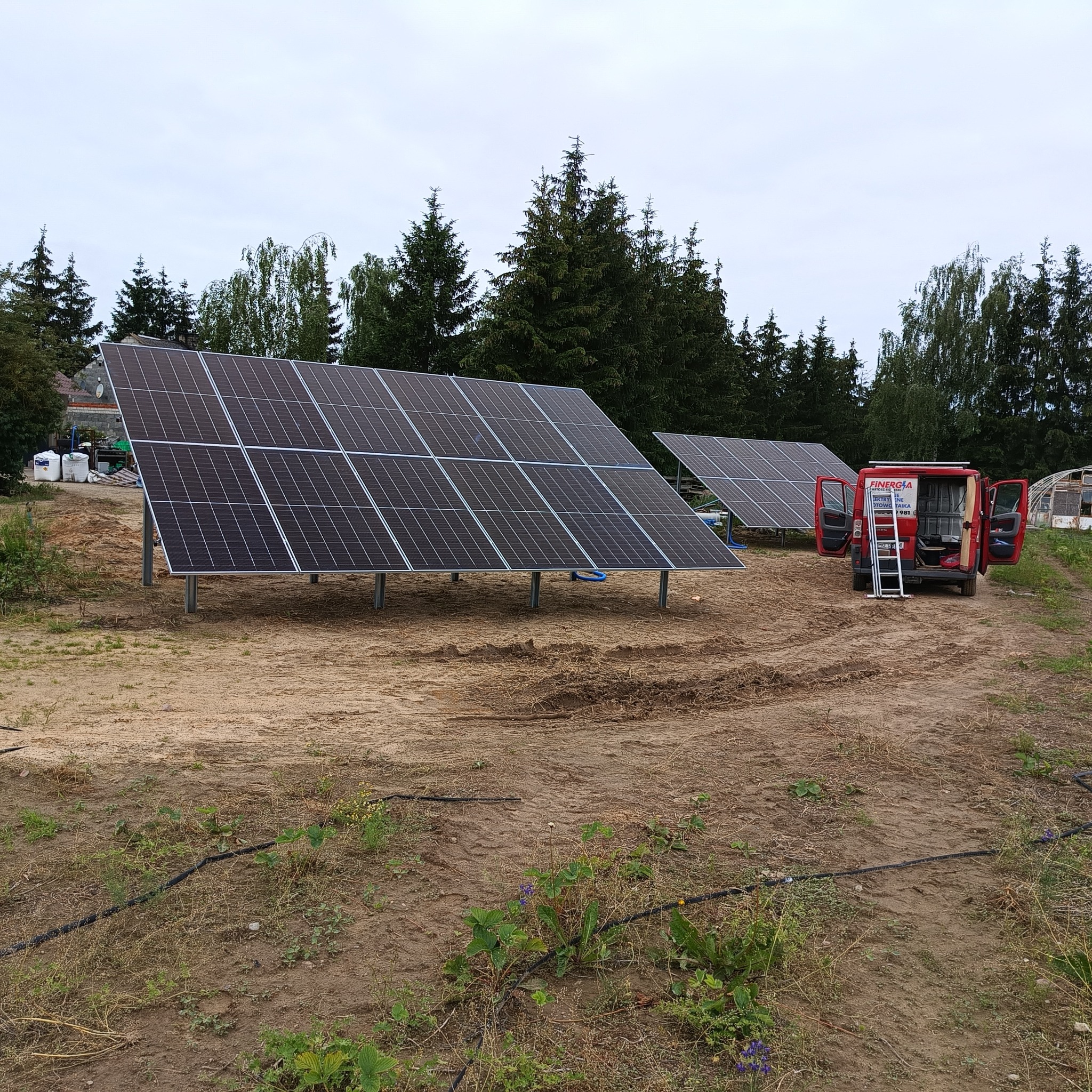 Panele słoneczne na metalowych stelażach, częściowo zainstalowane na piaszczystym terenie z widocznym transportowym samochodem dostawczym z otwartymi drzwiami i rozłożoną drabiną, w tle zieleń drzew.