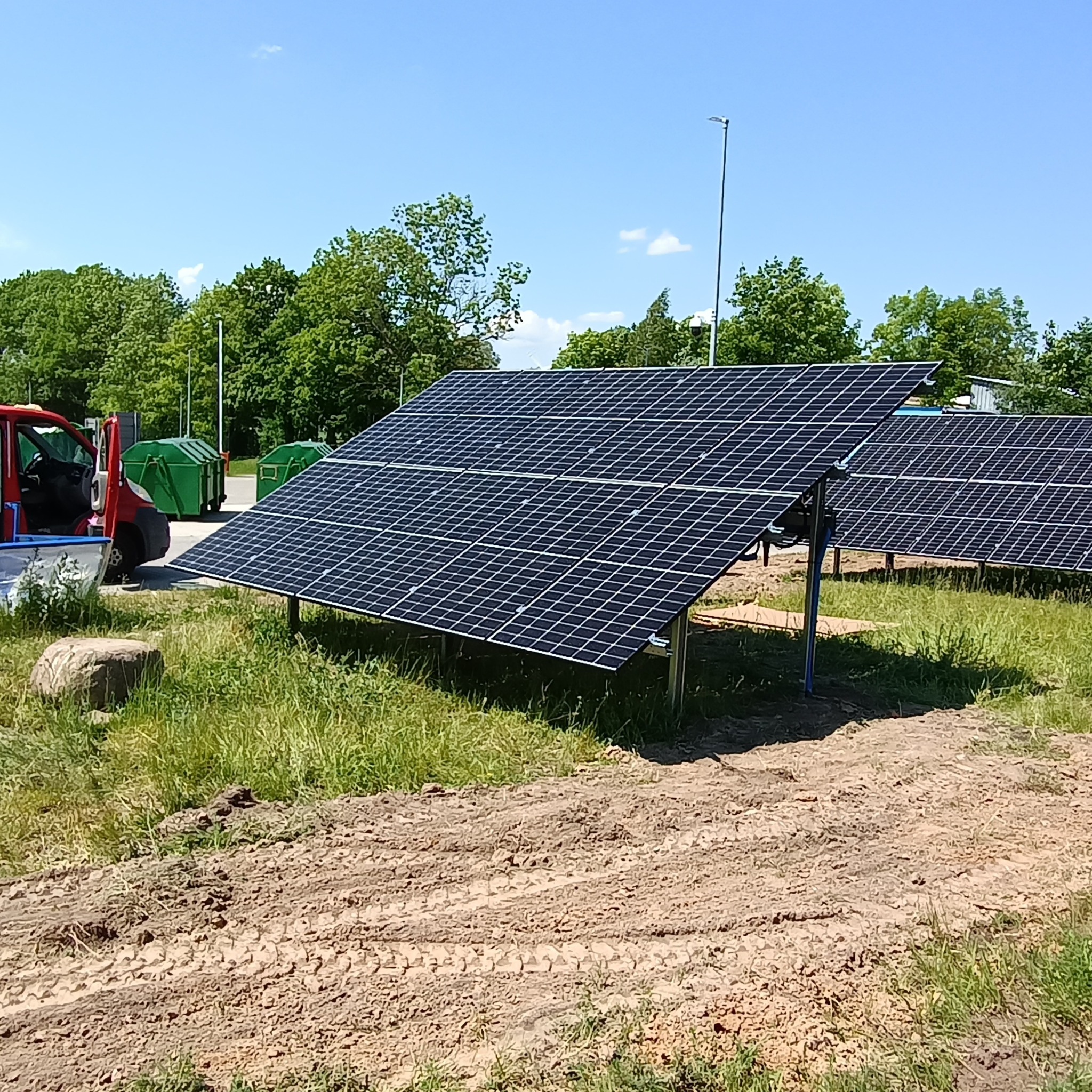 Panele słoneczne na metalowych stelażach na trawiastym terenie, w tle czerwony samochód dostawczy z otwartymi drzwiami i zielone kontenery na śmieci.