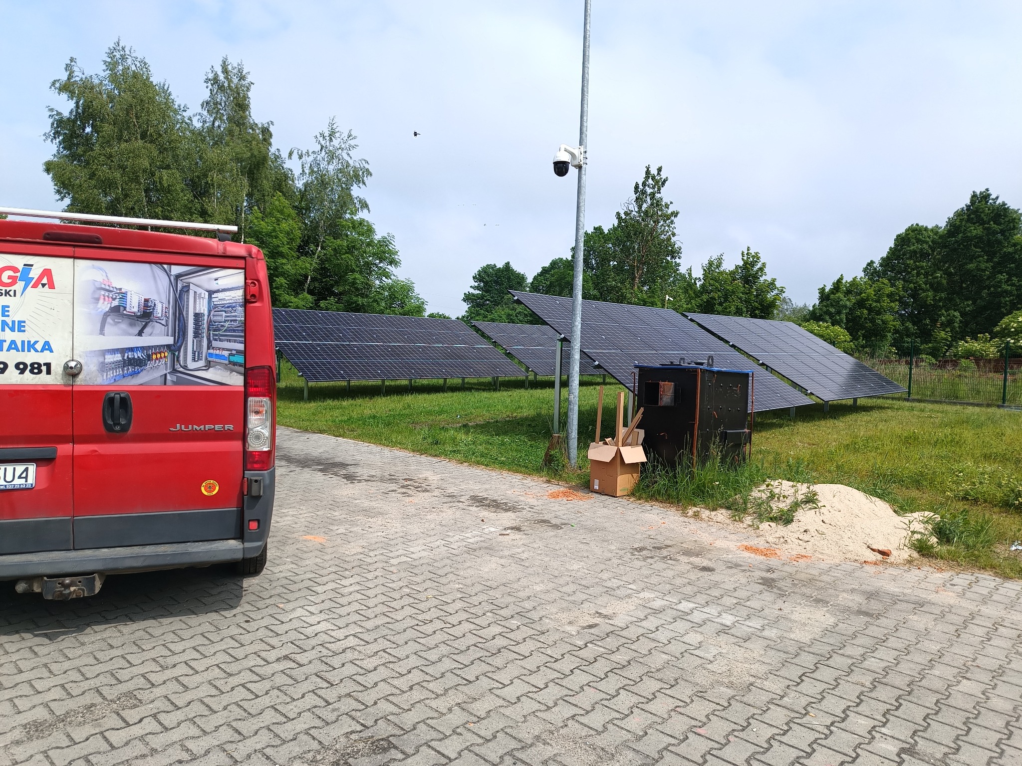 Panel słoneczny w rzędach na trawiastym terenie, widok z boku czerwonego samochodu dostawczego z reklamą firmy, kamera przemysłowa na słupie w tle, sterta piasku i karton na kostce brukowej.