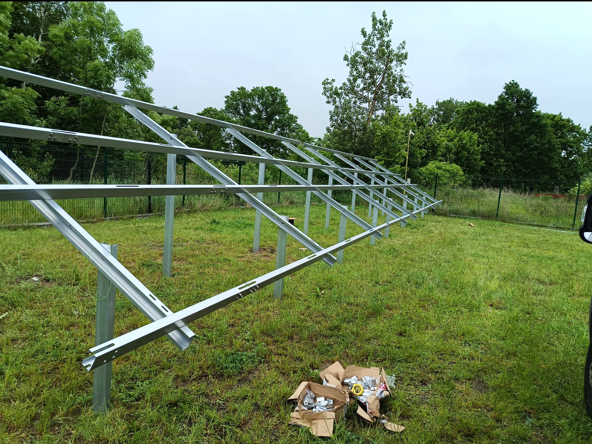 Konstrukcja wsporcza z profili stalowych pod panele fotowoltaiczne, montowana na trawniku, widoczny rozpakowany karton ze śrubami i elementami montażowymi.