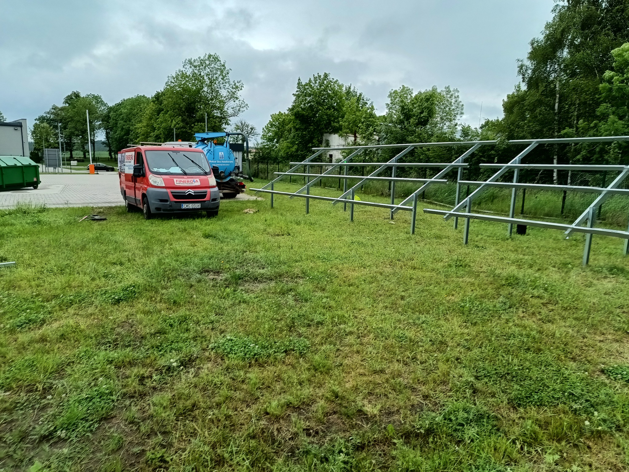 Montaż metalowej konstrukcji wsporczej pod panele słoneczne na trawiastym terenie, z czerwonym samochodem dostawczym firmy na drugim planie.