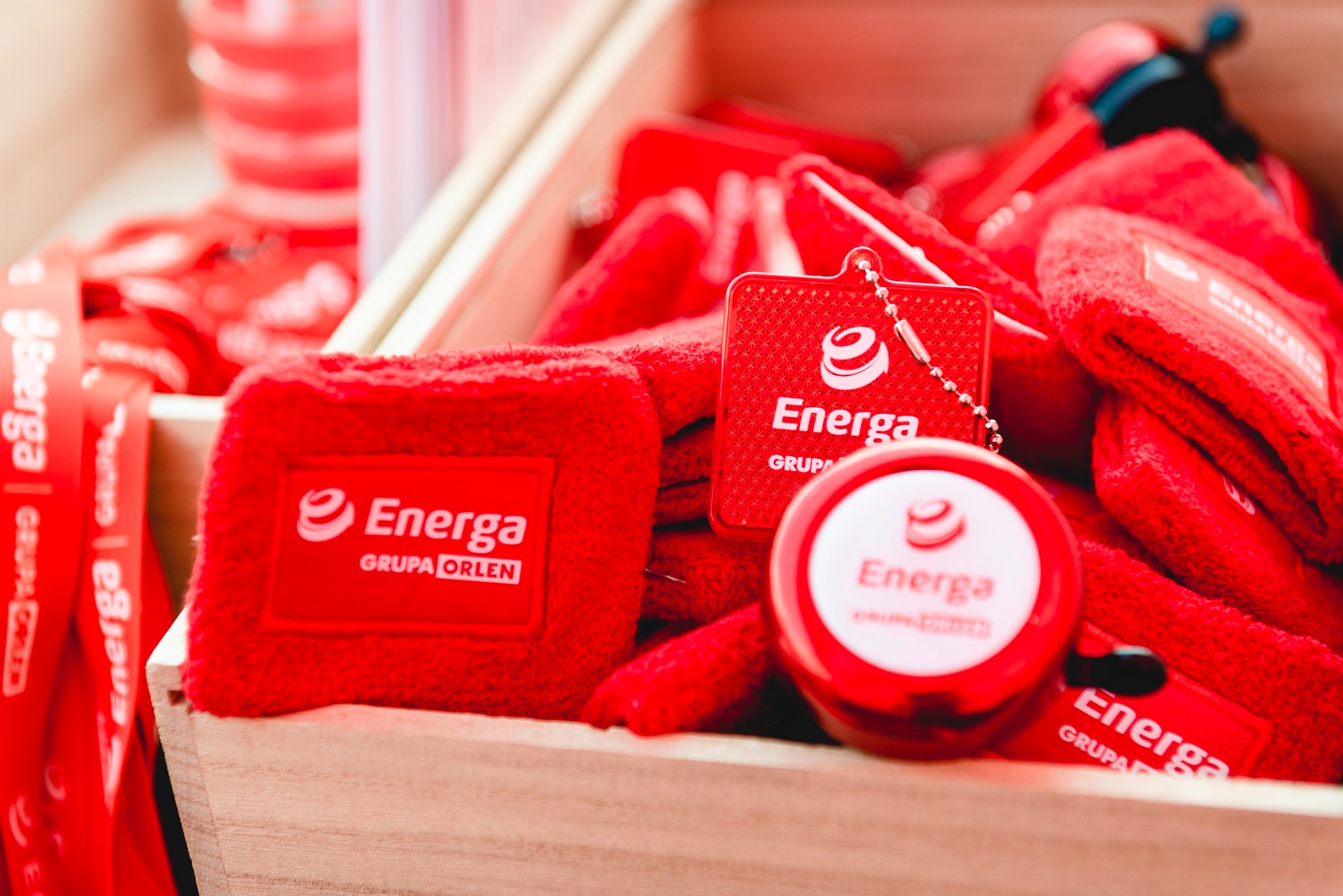 Czerwone gadżety promocyjne firmy Energa Grupa Orlen w drewnianej skrzynce: opaski na rękę, smycze, breloczek i miarka zwijana.