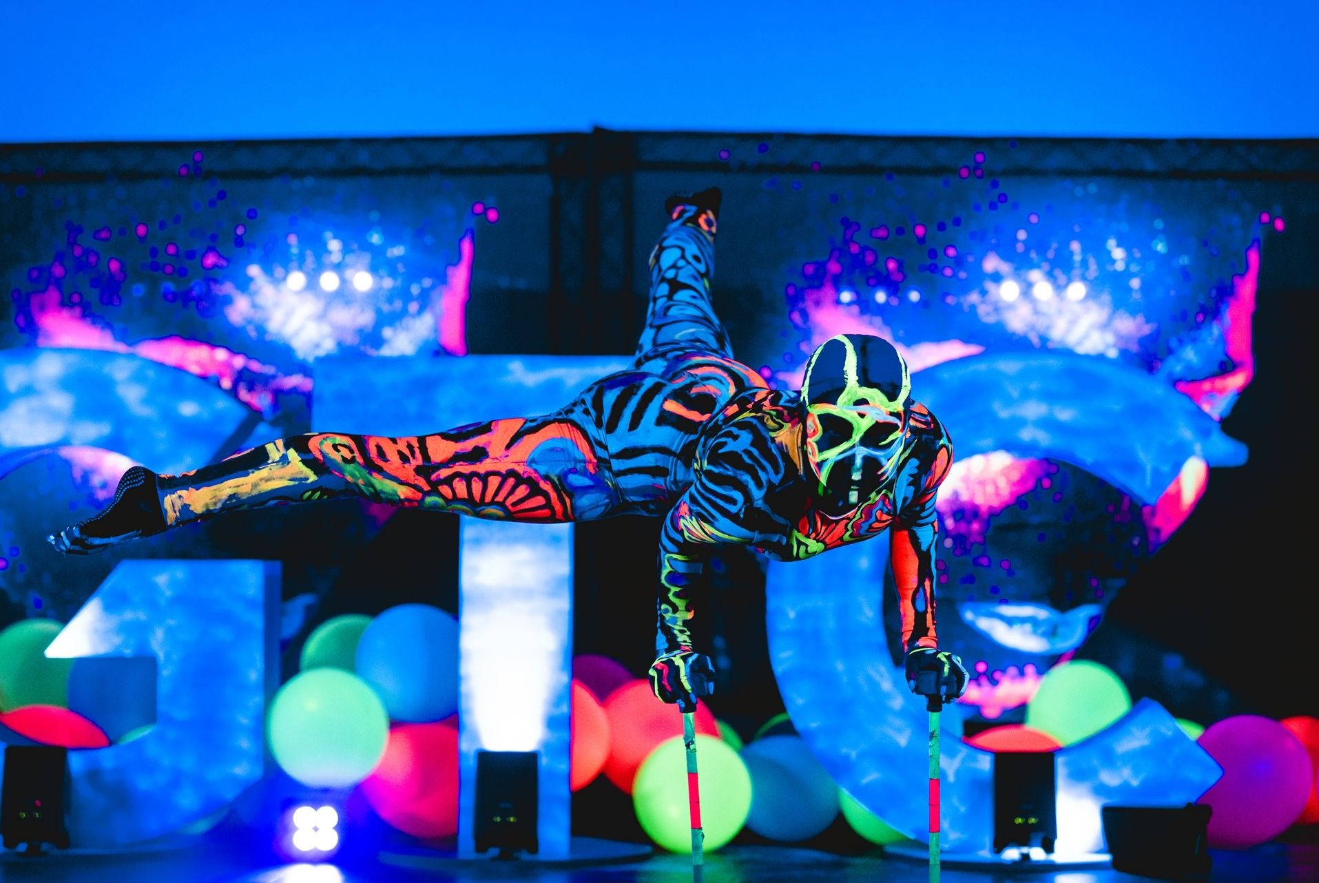 Akrobata w neonowym kostiumie, masce i na szczudłach, w pozycji horyzontalnej na tle podświetlonej sceny z balonami i dekoracjami.