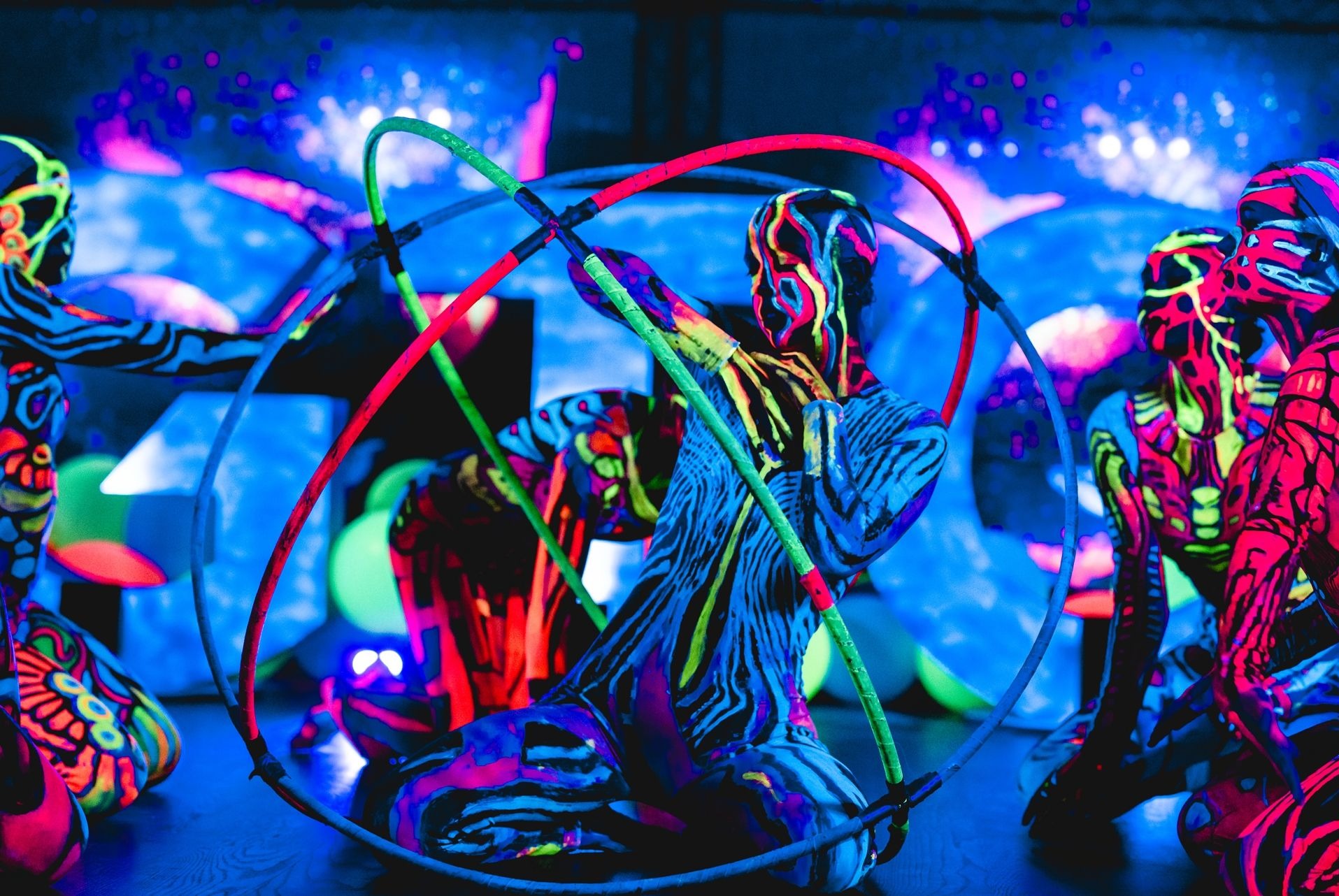 Grupa artystów z pomalowanymi ciałami w neonowych barwach, prezentujących performance z rekwizytami świetlnymi na ciemnej scenie z niebieskim oświetleniem.