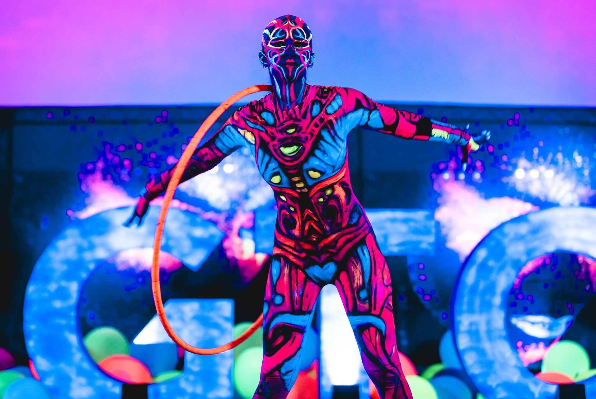 Osoba z pomalowanym ciałem w jaskrawych, neonowych barwach, trzymająca pomarańczowe koło, na tle dekoracji z podświetlanych liter i balonów.