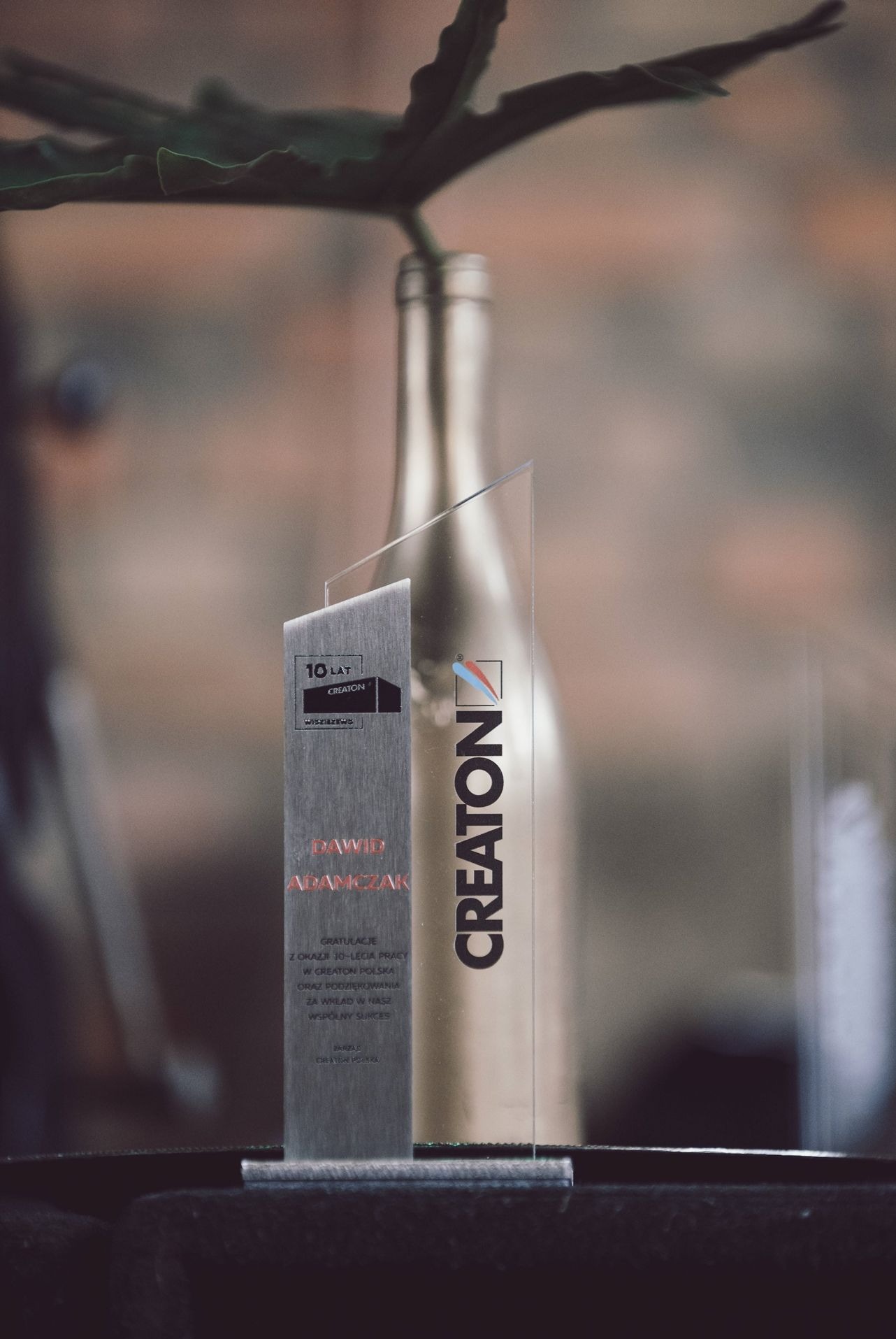 Szklana statuetka z grawerem 'CREATON' i napisem '10 lat' dla Dawida Adamczaka, stojąca na ciemnej powierzchni, z rozmytym tłem w odcieniach brązu i zieleni.