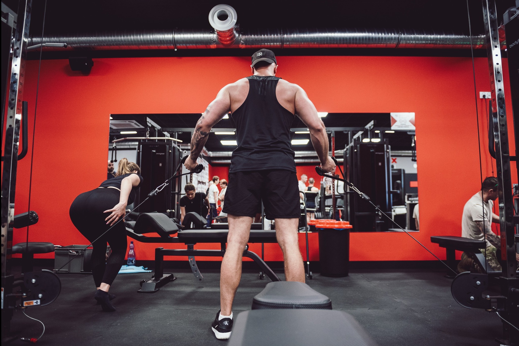 Mężczyzna z tatuażami na ramionach ćwiczy na maszynie wyciągowej w siłowni z czerwonymi ścianami i lustrami, widoczni inni ćwiczący.