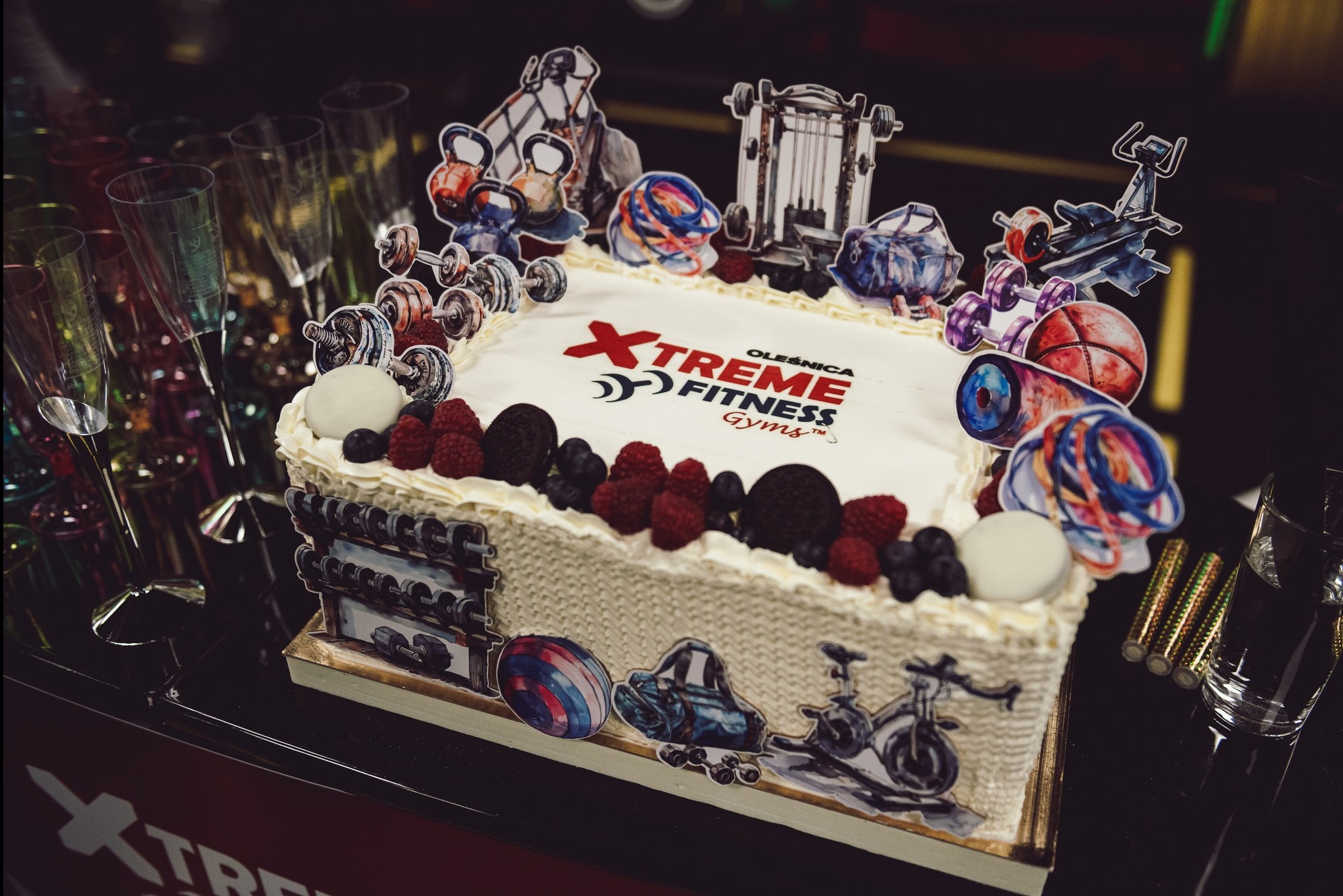 Prostokątny tort z logo 'Xtreme Fitness Gyms', ozdobiony owocami, ciasteczkami Oreo i papierowymi dekoracjami przedstawiającymi sprzęt do ćwiczeń, stoi na czarnym stole obok szklanek.