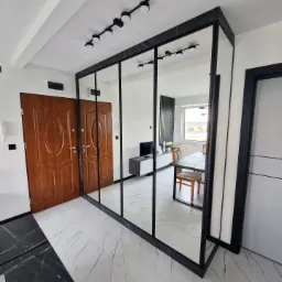 Przestronny przedpokój z dużą szafą wnękową z lustrzanymi drzwiami przesuwnymi, czarne ramy i marmurowy wzór na obrzeżach, odbicie wnętrza mieszkania w lustrach.