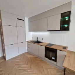 Nowoczesna kuchnia z szarymi frontami, drewnianym blatem, oświetleniem LED w szafce wiszącej i parkietem w jodełkę.