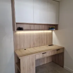 Biurko na wymiar z górnymi szafkami w kolorze białym i drewnopodobnym, z oświetleniem LED podszafkowym, półkami na książki i akcesoria, widoczne dwie książki i dwa aparaty fotograficzne, szare...