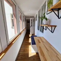 Wąski, jasny korytarz z drewnianą podłogą i blatem, zabudowany wzdłuż okna z siedziskiem z niebieską tapicerką, białe ściany z półkami z roślinami.