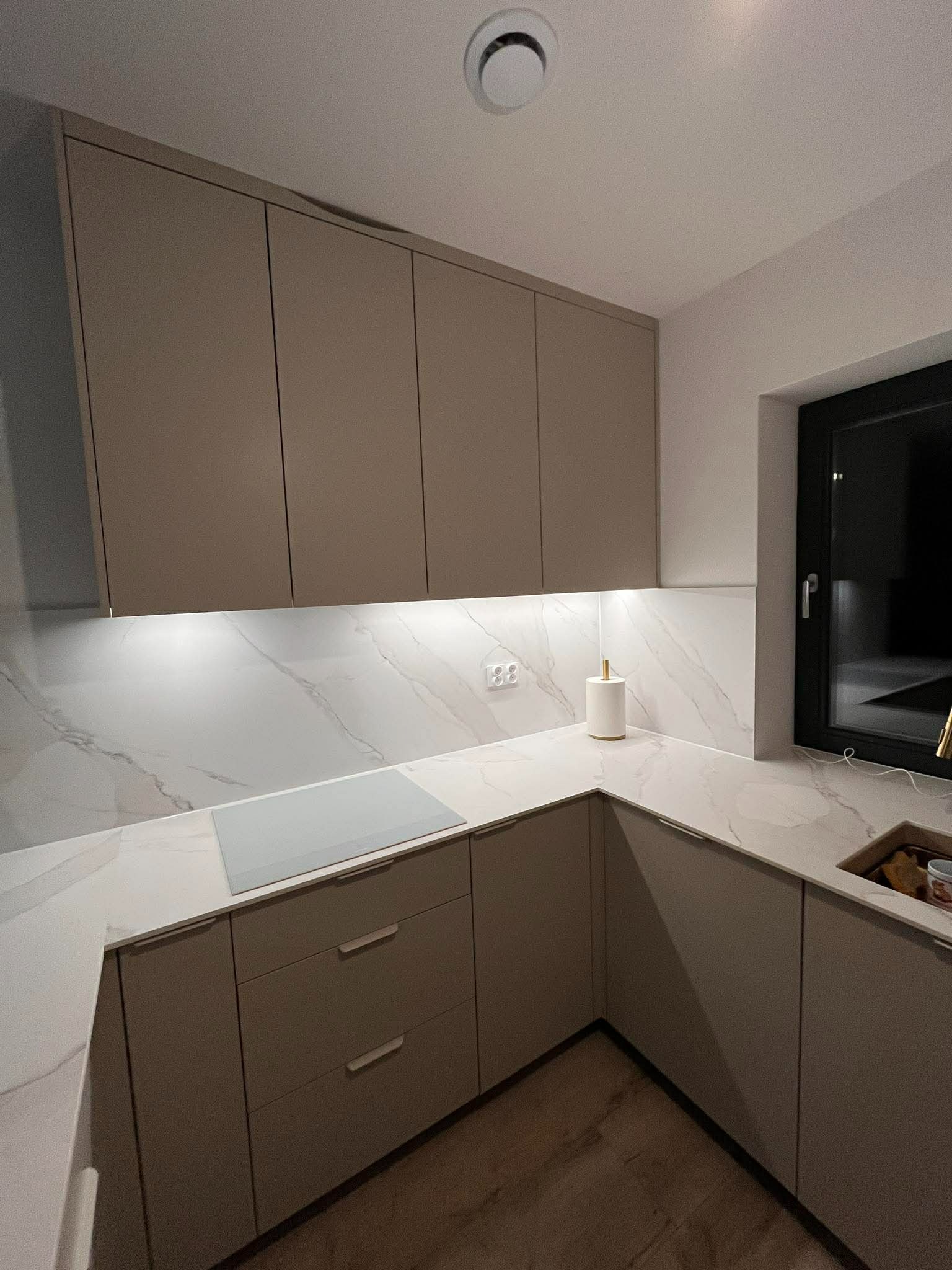 Beżowa kuchnia z marmurowym blatem i oświetleniem LED pod szafkami wiszącymi. Minimalistyczny design, płyta indukcyjna wtopiona w blat. Nowoczesne wykończenie.