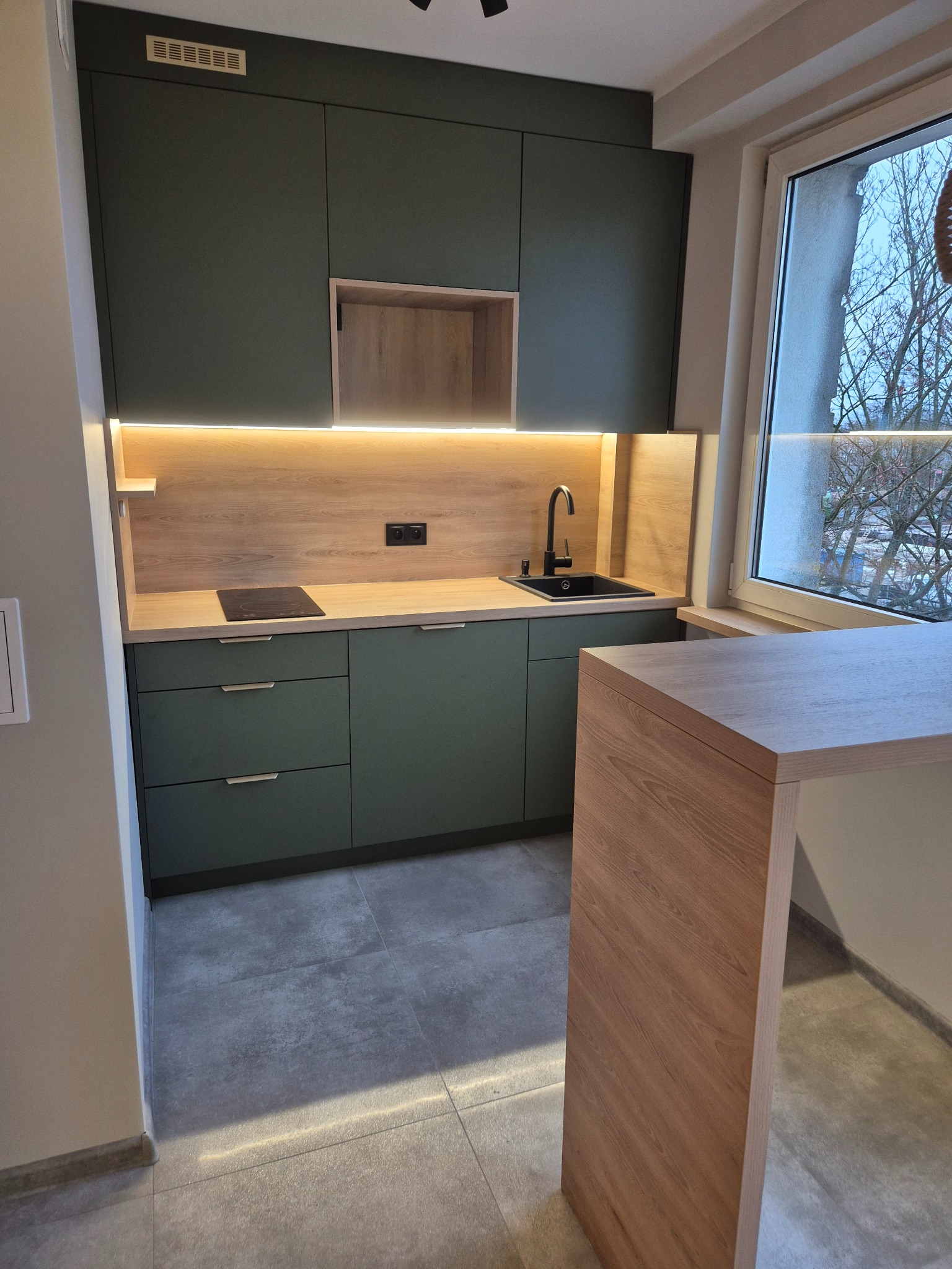 Nowoczesna kuchnia z zielonymi szafkami, drewnianym blatem i oświetleniem LED. Minimalistyczny design z czarną armaturą i płytą indukcyjną. Widok na okno.