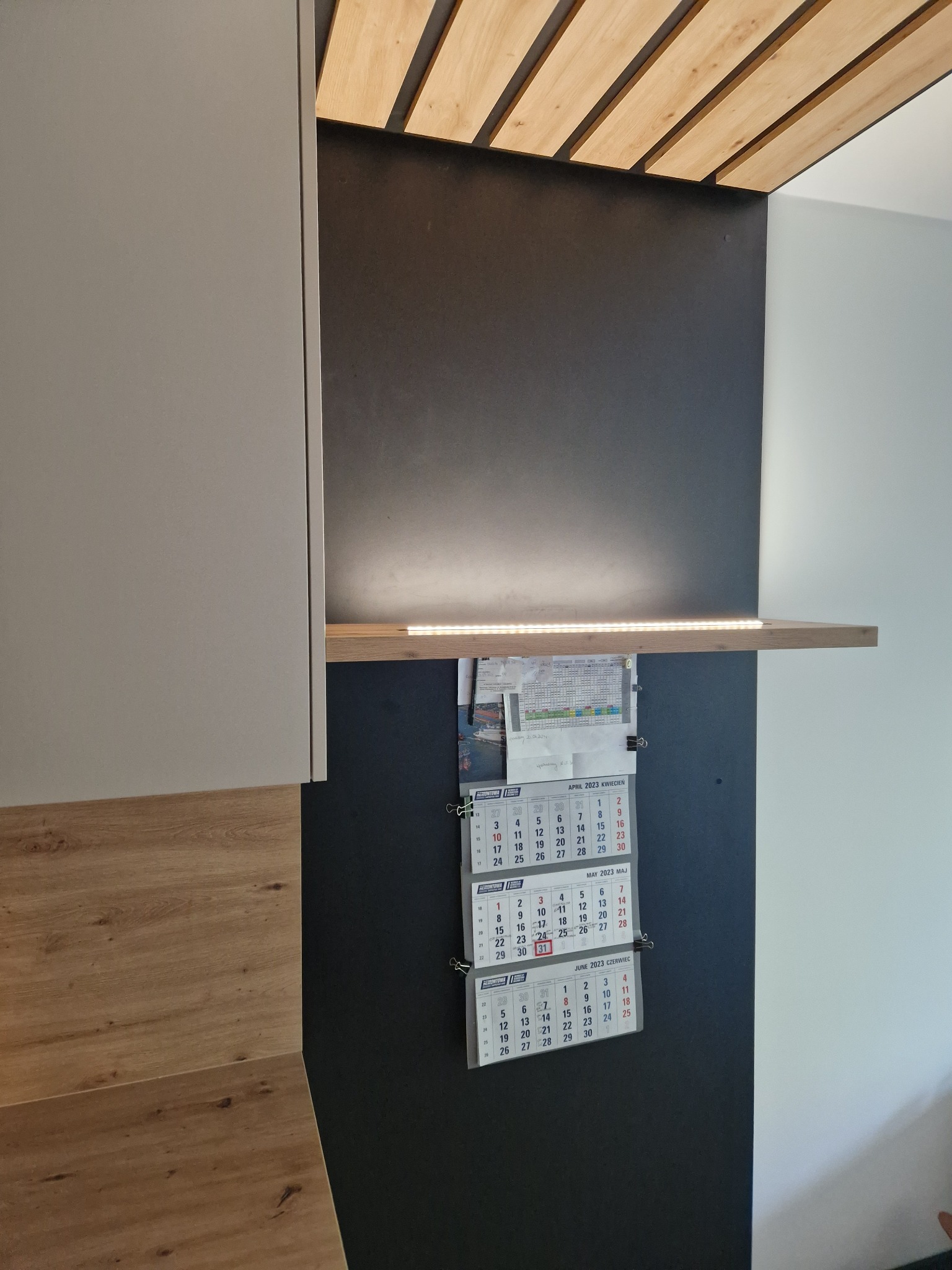 Fragment zabudowy kuchennej z drewnianym blatem i górną szafką w jasnym odcieniu, podświetlona listwą LED, z czarną ścianą i drewnianym panelem sufitowym. Na ścianie wiszą trzy kalendarze.