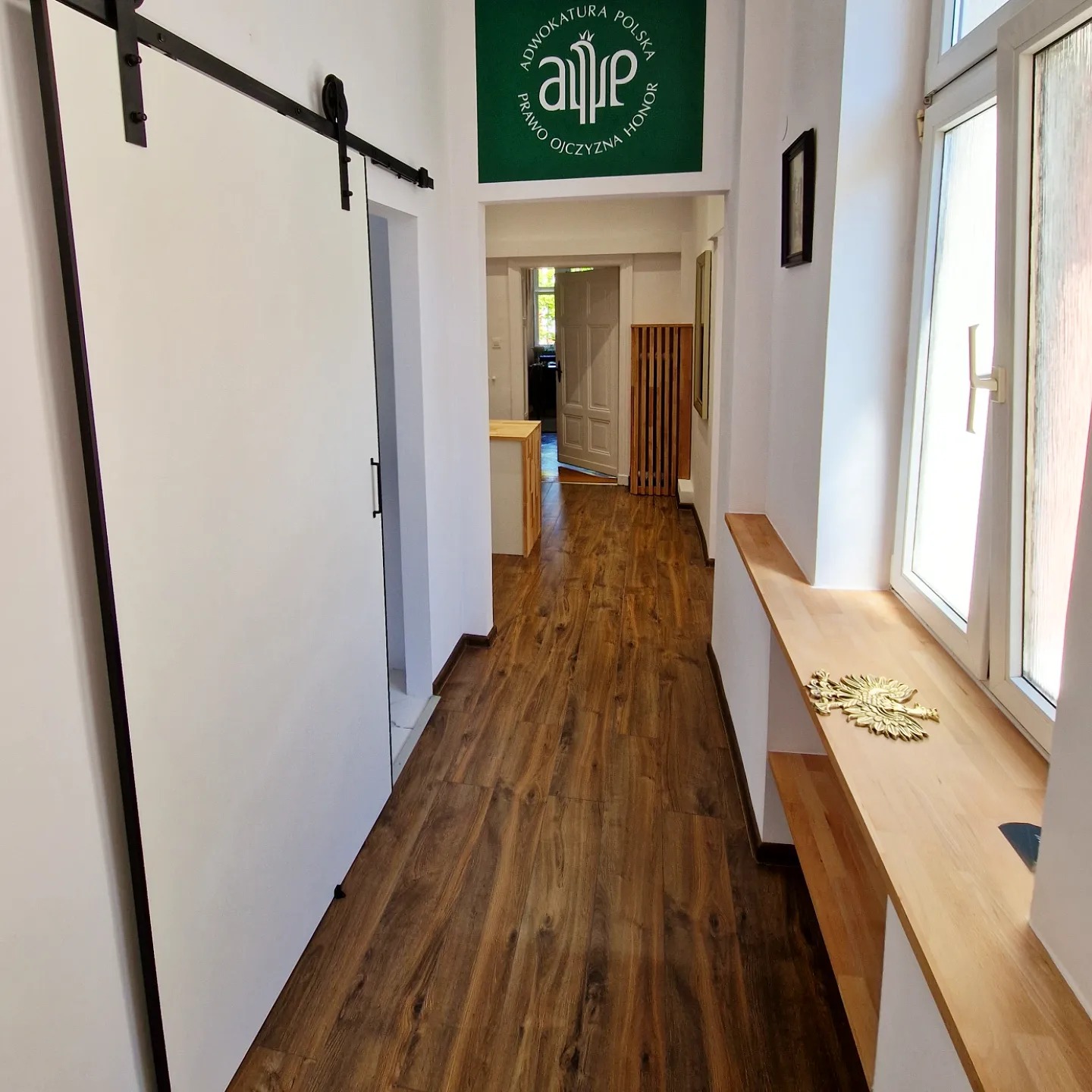 Wnętrze korytarza z przesuwnymi drzwiami na czarnych szynach, drewnianą podłogą, oknem z drewnianym parapetem i dekoracją w kształcie słońca, widok na dalsze pomieszczenia.