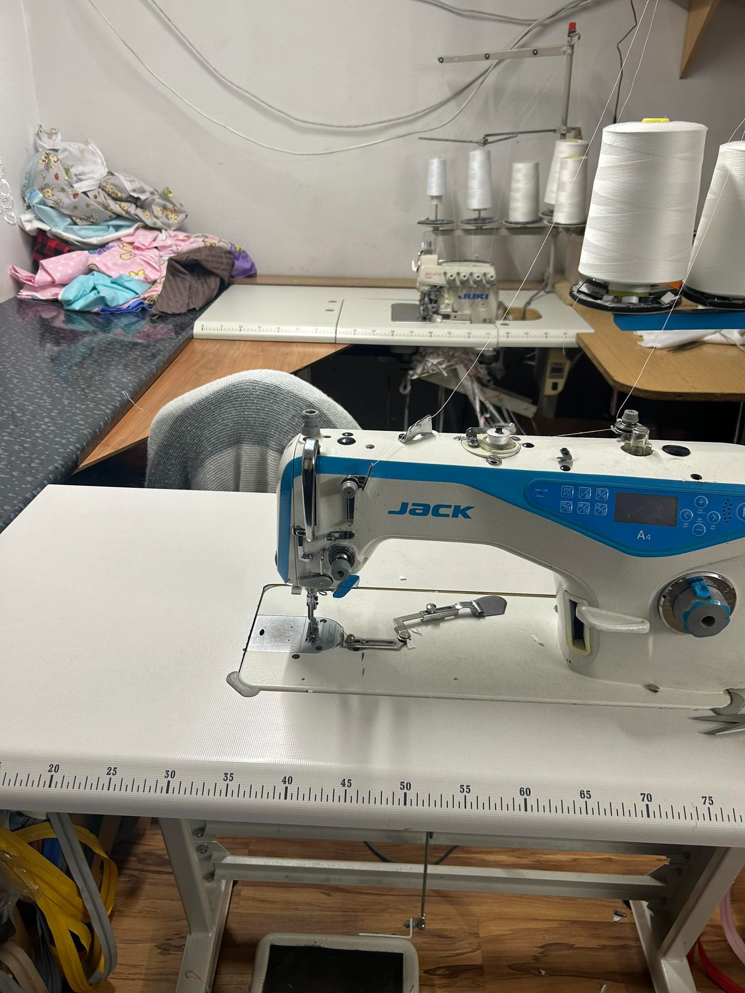 Profesjonalna maszyna do szycia marki Jack A4 na stole z miarką, w tle inne maszyny, szpule nici i stos tkanin.