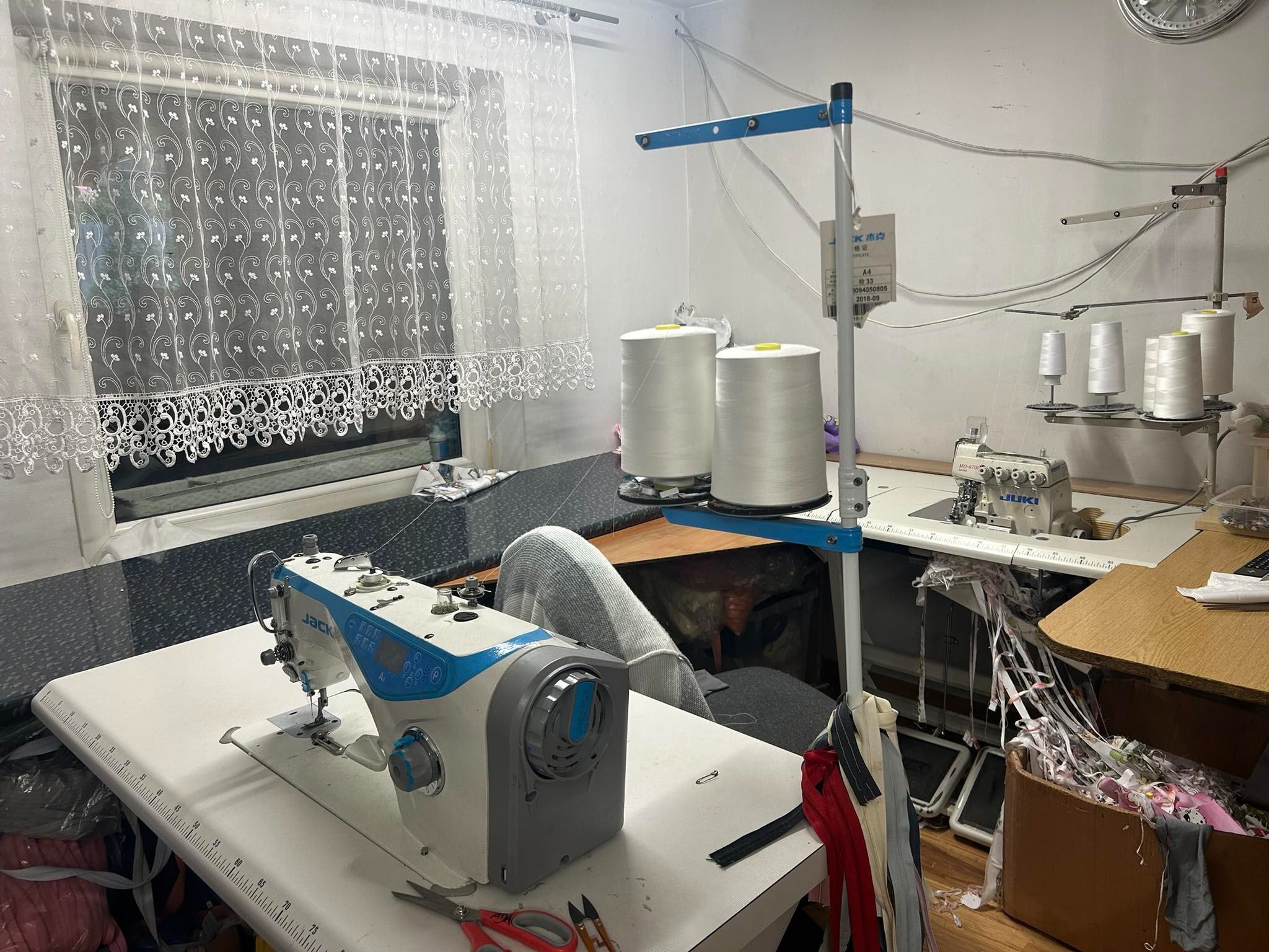 Stanowisko pracy w szwalni: maszyna do szycia Jack z niebieskimi akcentami na stole z miarką, obok dwie szpule białej nici, w tle okno z koronkową firanką i druga maszyna Juki.