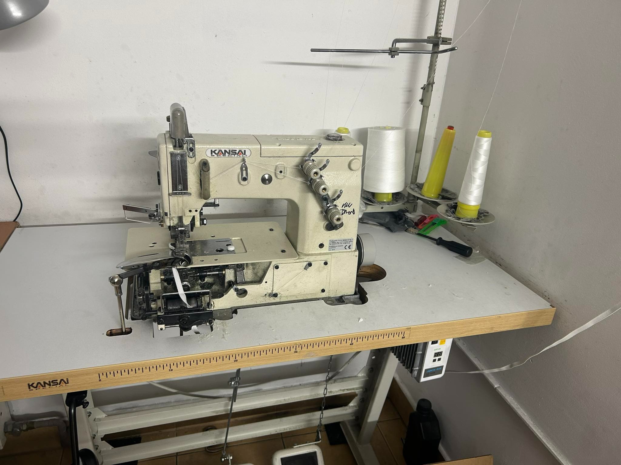 Przemysłowa maszyna do szycia marki Kansai, ustawiona na stole z miarką, z widocznymi szpulkami nici i elementami konstrukcyjnymi, w warsztacie krawieckim.