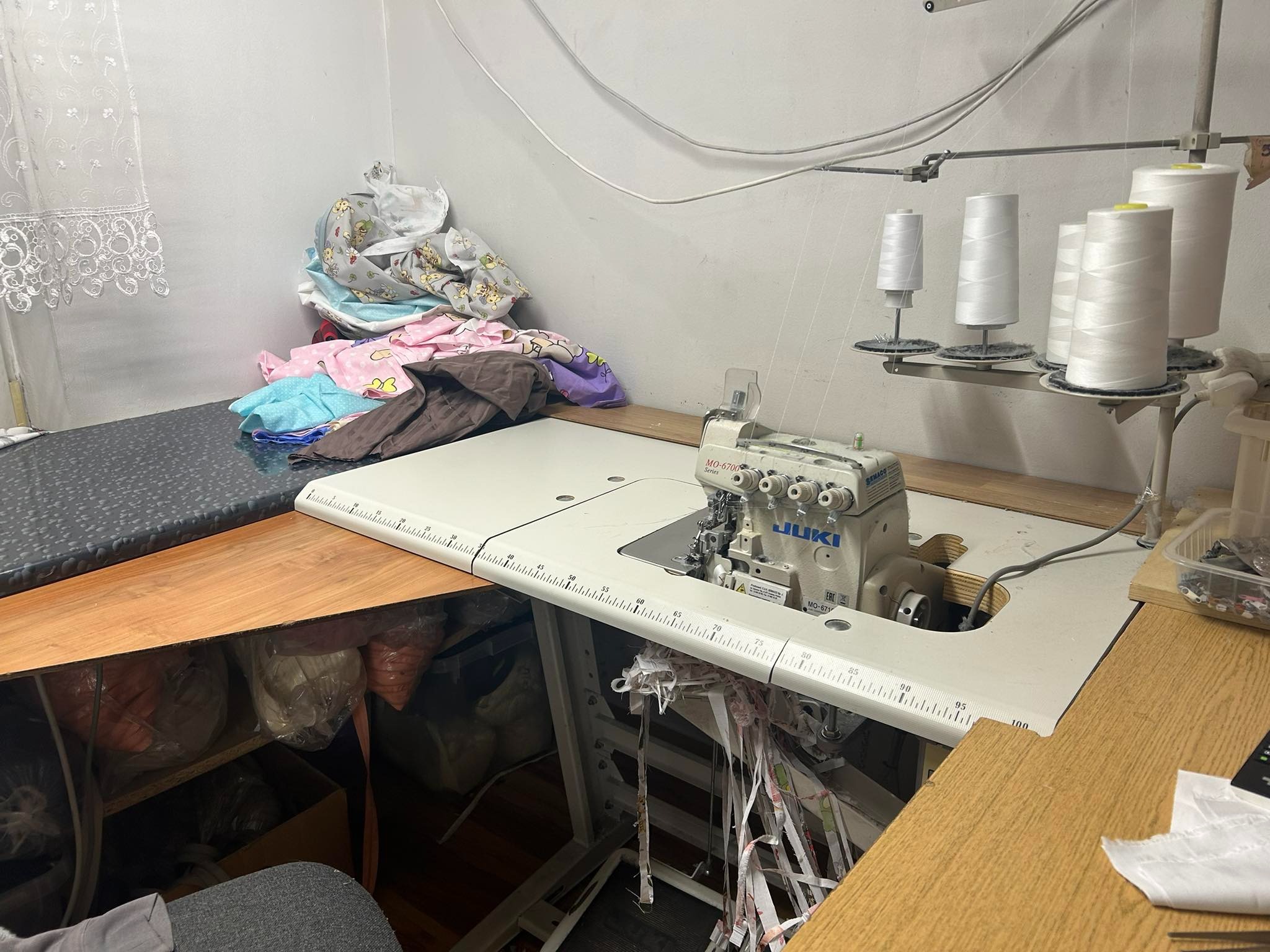 Przemysłowa maszyna do szycia JUKI MO-6714 na stole roboczym z miarką, w tle stos materiałów z wzorami, oraz szpulki z białymi nićmi na stojaku.