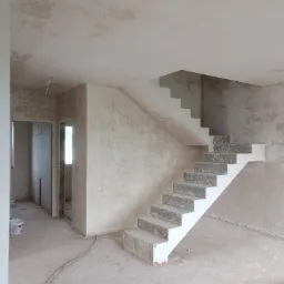 Wnętrze w stanie deweloperskim ze świeżo nałożonymi tynkami na ścianach i suficie oraz surowymi schodami betonowymi z białą obudową, widoczne kable na podłodze i otwarte przejścia do innych...