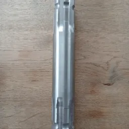 Metalowy wałek z rowkami i wycięciami, leżący na drewnianej powierzchni, prawdopodobnie po obróbce skrawaniem.