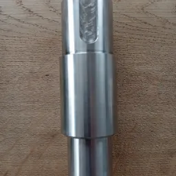 Metalowy element po obróbce, z widocznymi śladami spawania w wyfrezowanym rowku, leżący na drewnianej powierzchni.