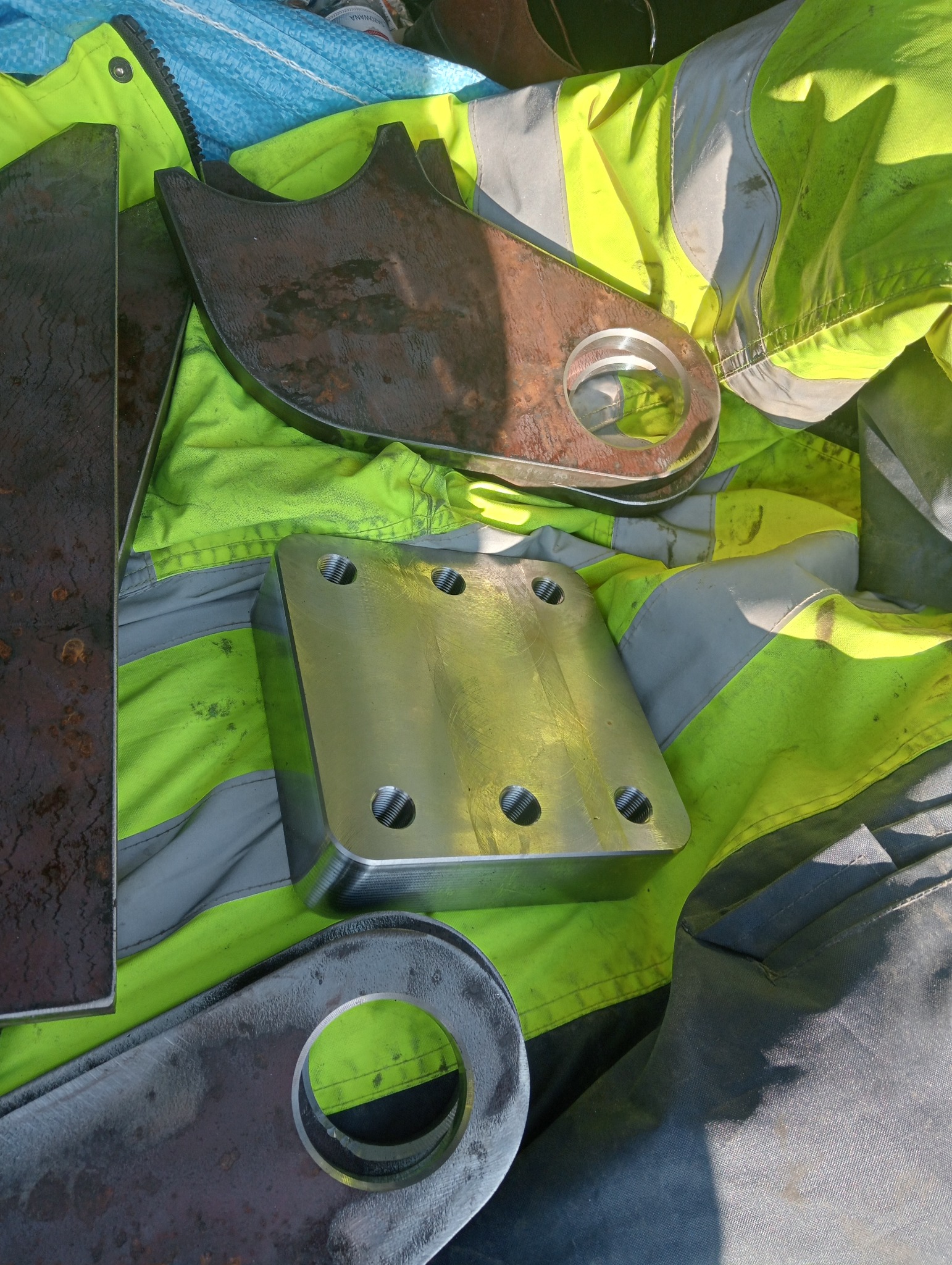 Różne metalowe elementy, w tym kwadratowa płyta z otworami gwintowanymi i elementy z wyciętymi okręgami, leżące na roboczej odzieży w kolorze neonowej zieleni i szarości.