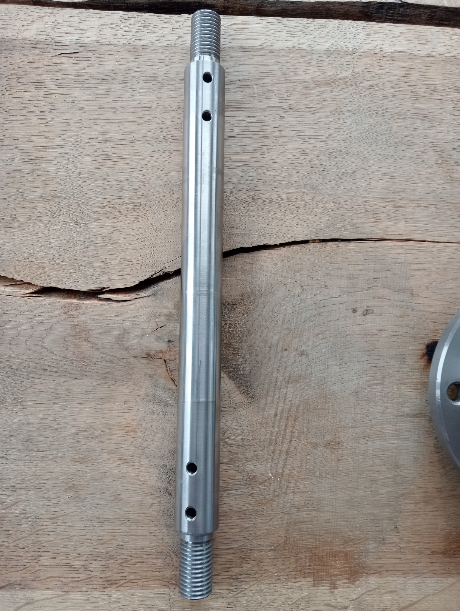 Metalowy, gwintowany wałek z otworami, leżący na drewnianej powierzchni z widocznymi słojami.