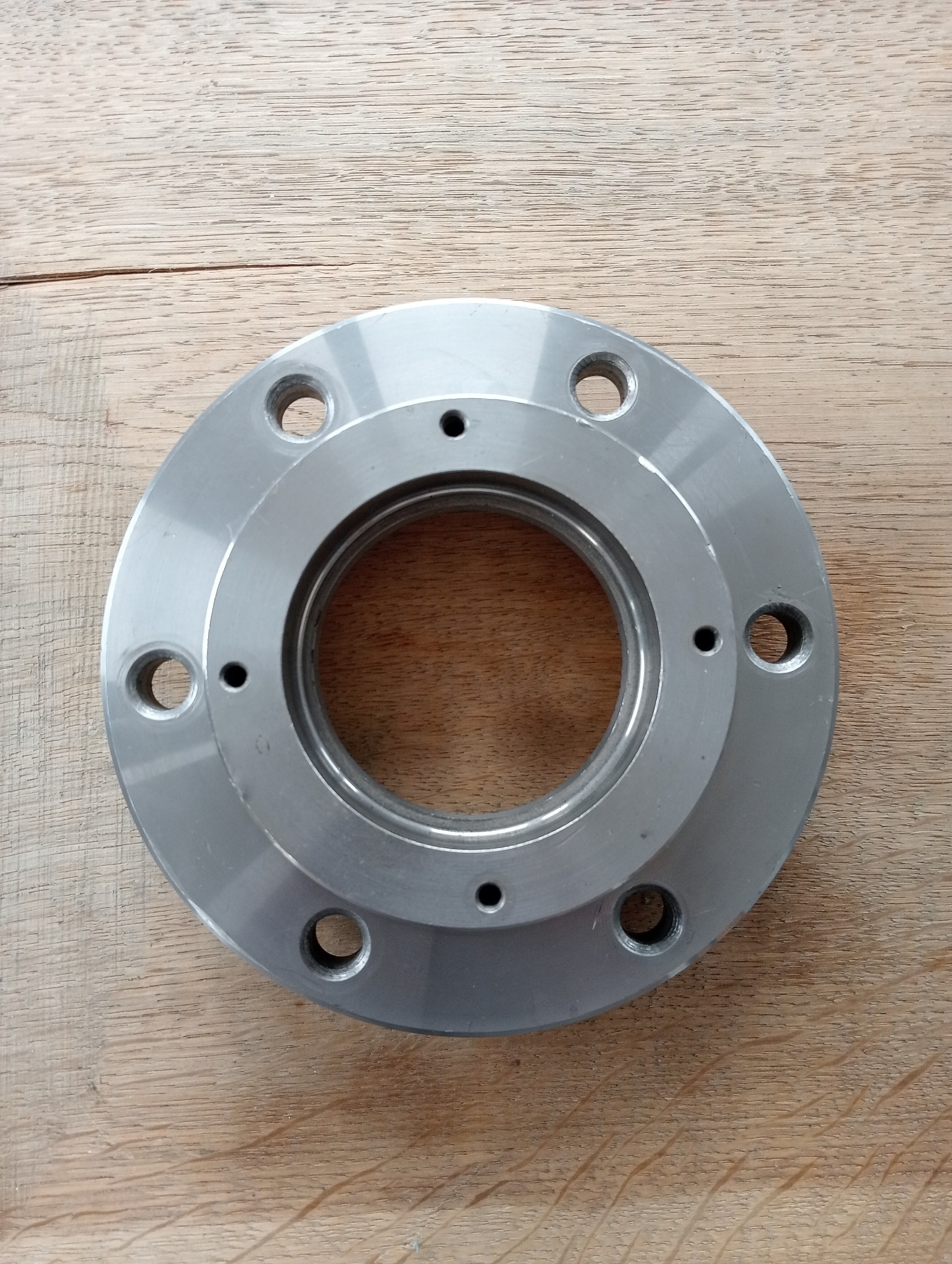 Szlifowany, okrągły element metalowy z otworami na śruby, leżący na drewnianej powierzchni.