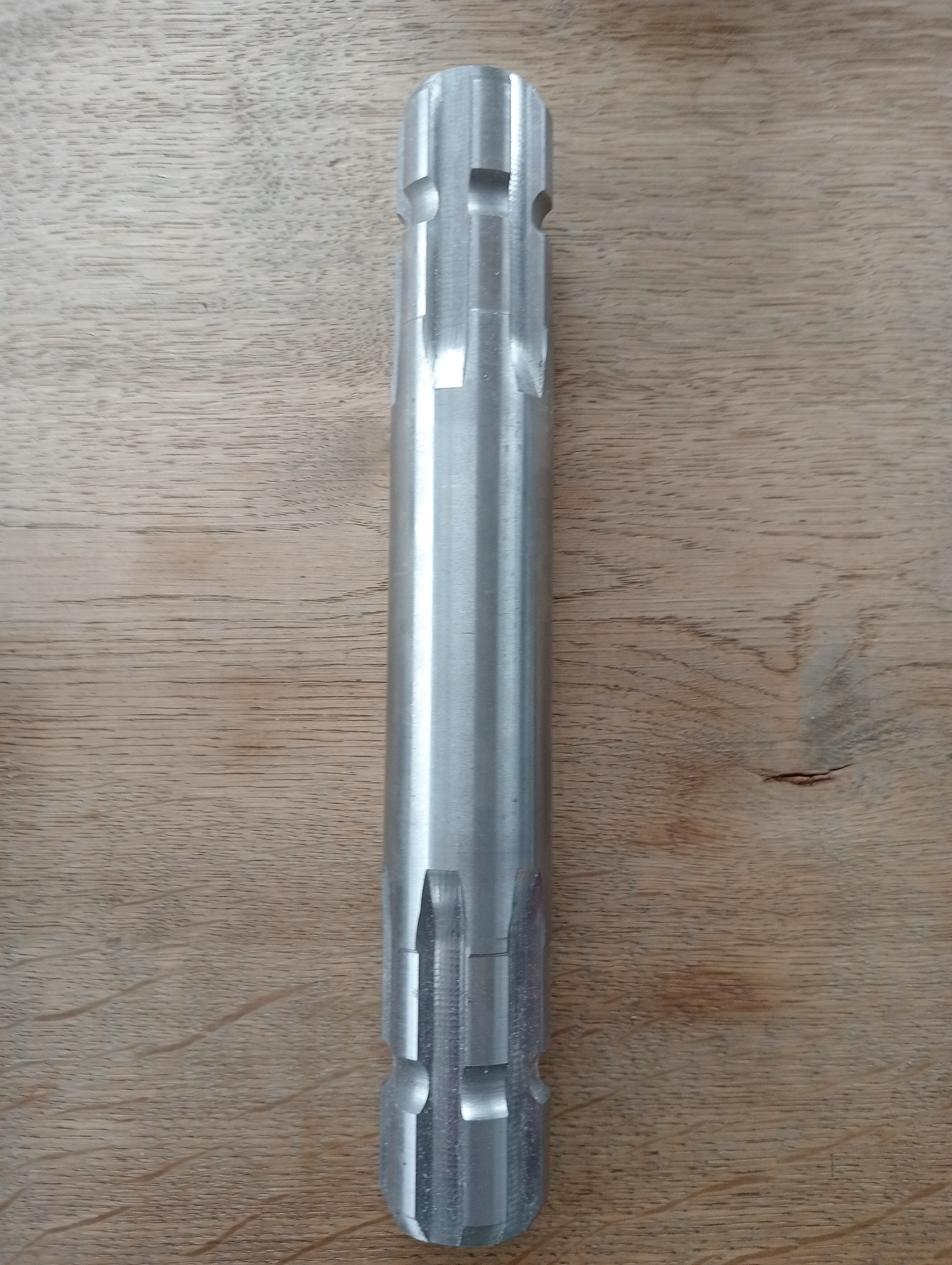 Metalowy wałek z rowkami i wycięciami, leżący na drewnianej powierzchni, prawdopodobnie po obróbce skrawaniem.