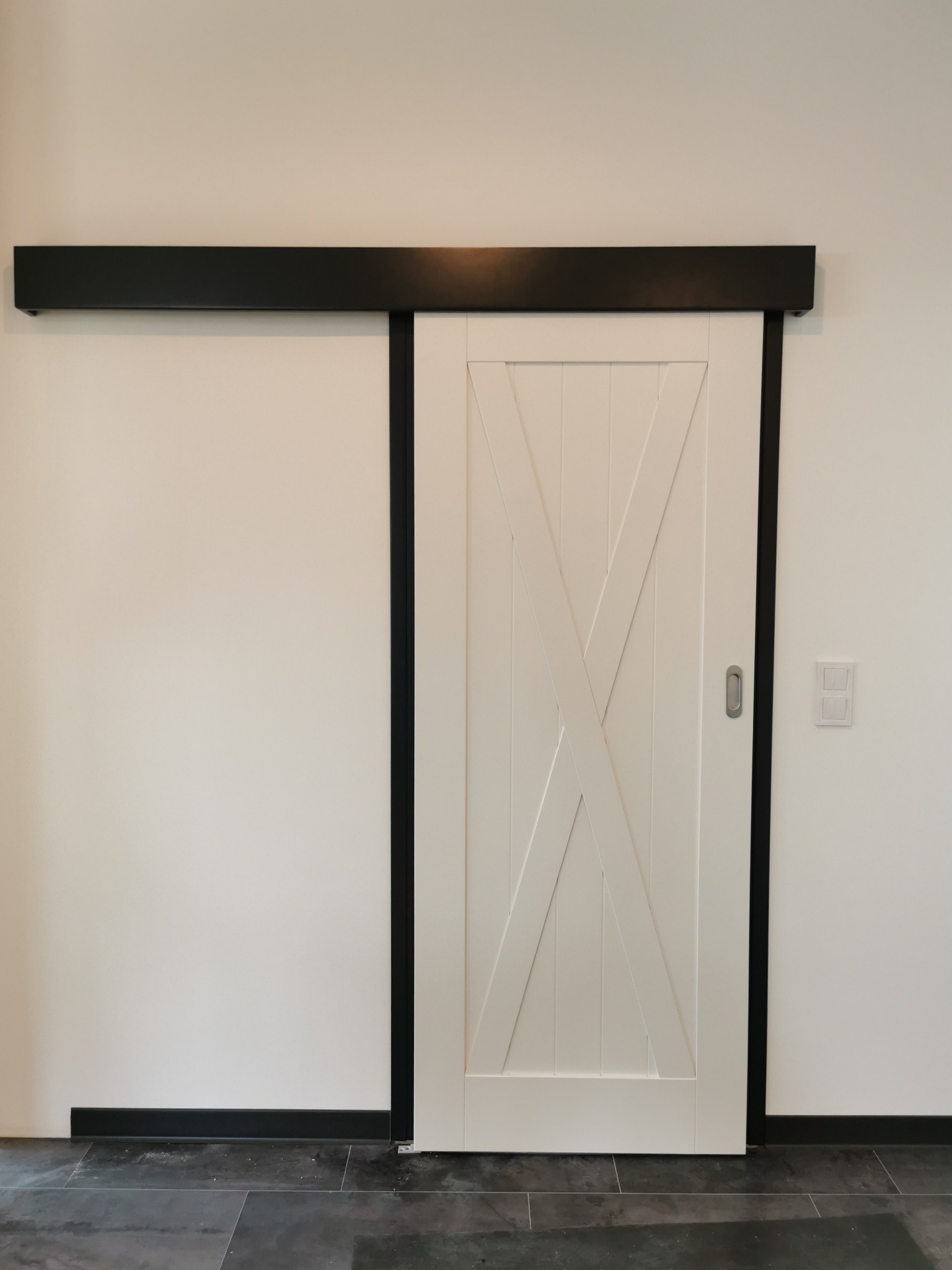 Białe drzwi przesuwne w stylu stodoły z czarną szyną i ramą, widoczne na tle białej ściany z szarymi płytkami podłogowymi.