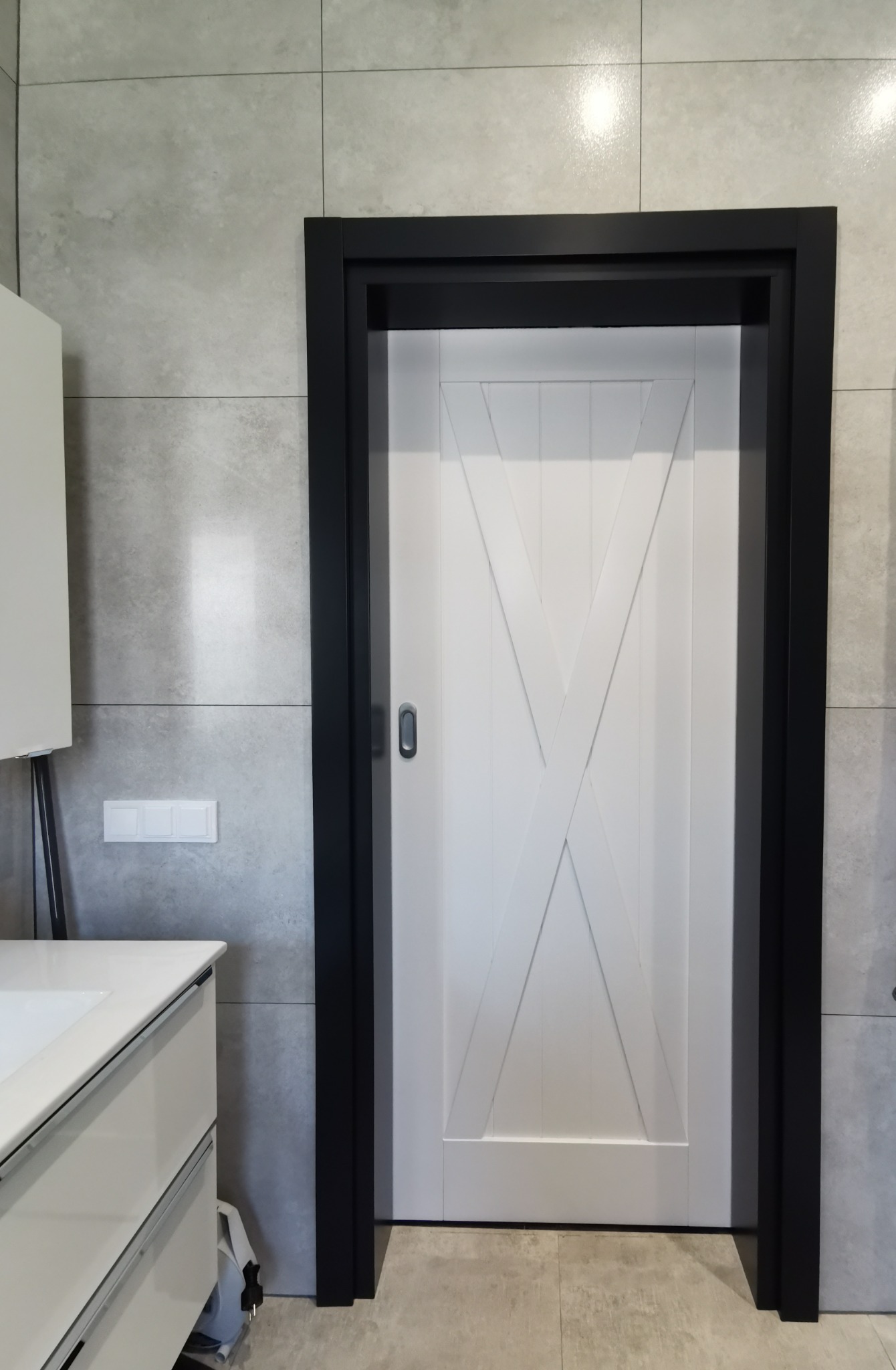 Białe drzwi wewnętrzne z czarną ościeżnicą i dekoracyjnym krzyżem, zamontowane w łazience z szarymi płytkami i białą szafką umywalkową.