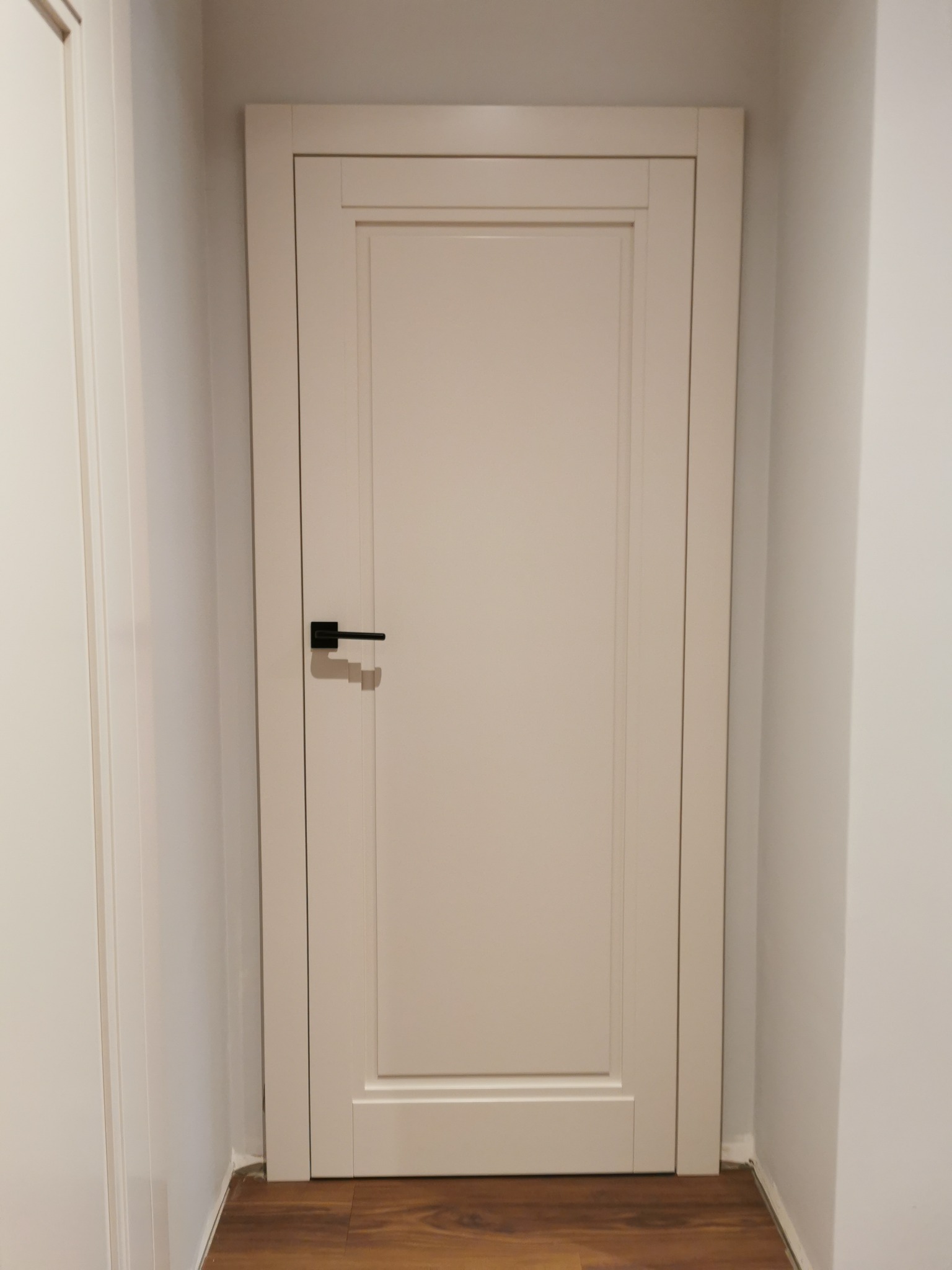 Zamknięte, kremowe drzwi wewnętrzne z czarną klamką na tle białej ściany i drewnianej podłogi.